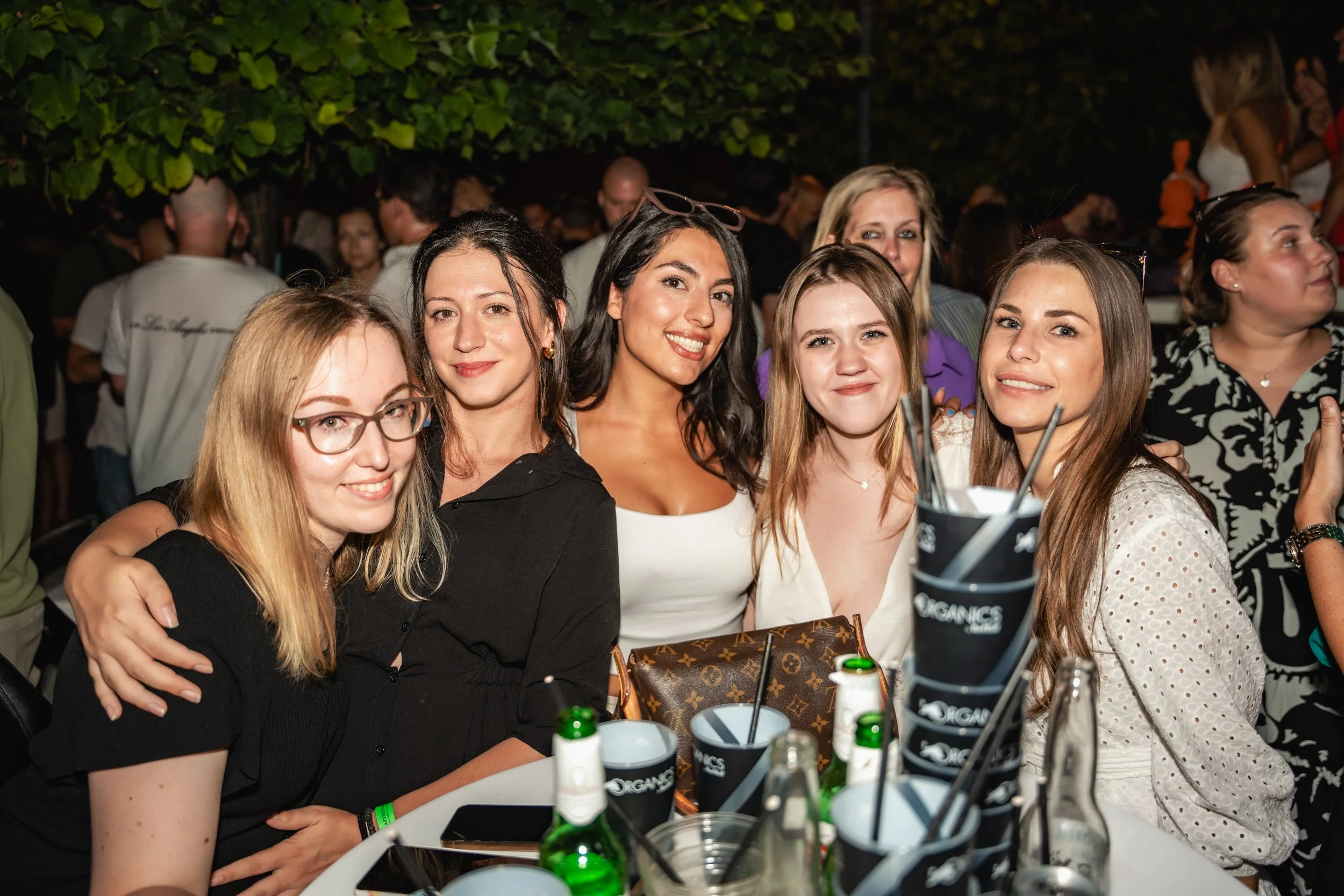 Group of young women smiling at a party or gathering outdoors at night, with drinks and bottles on the table in front of them.