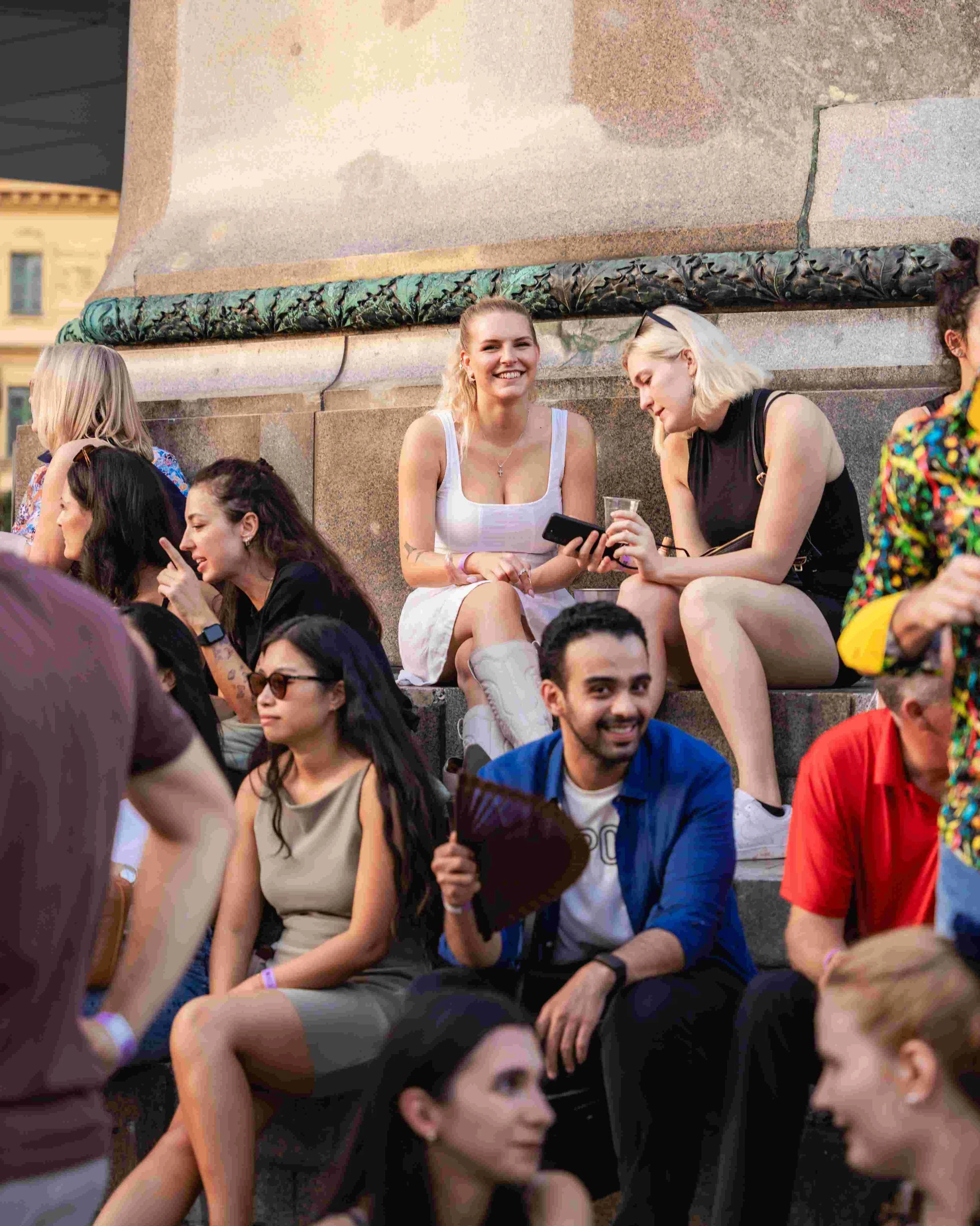 A group of young people sitting on steps at an outdoor event, smiling and chatting, with some holding smartphones and drinks.