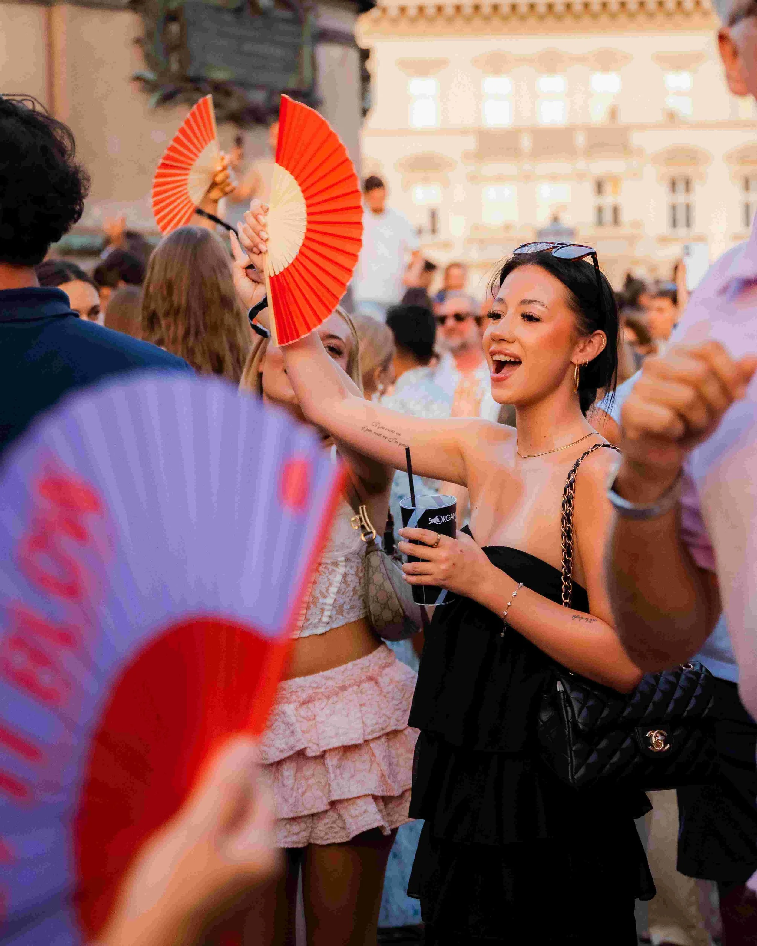 People gathered at an outdoor event with a woman smiling and holding a drink. She wears sunglasses on her head, a black outfit with gold chain straps, and has tattoos on her arm. Other people are in the background, some dancing and holding fans.