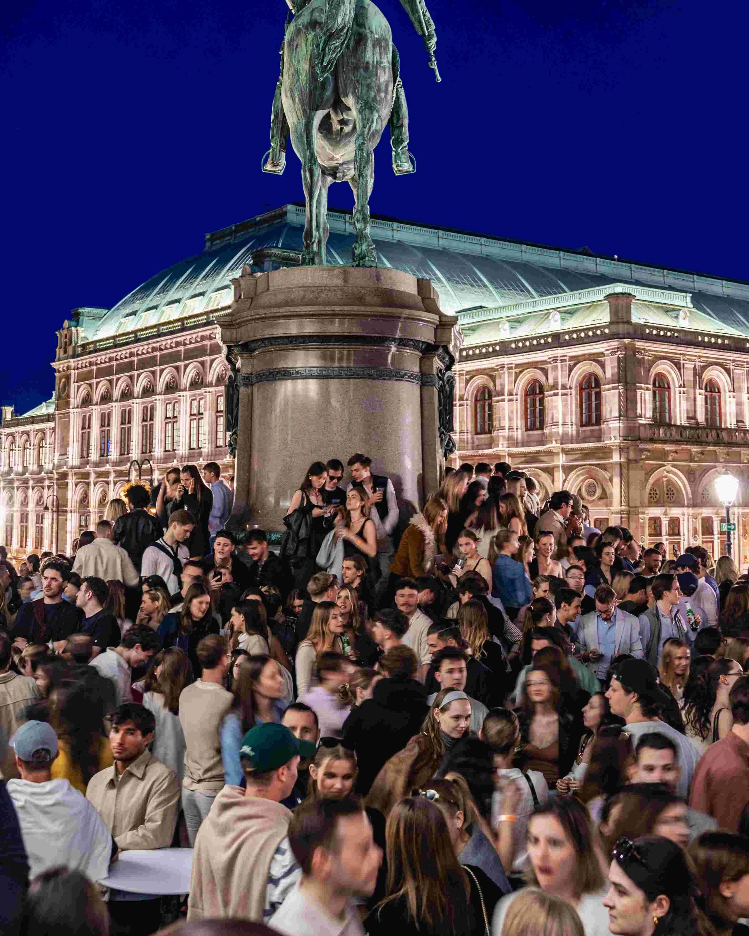 Crowd of people gathered in front of a historic building with a large bronze statue of a horse and rider on a pedestal, illuminated at night.