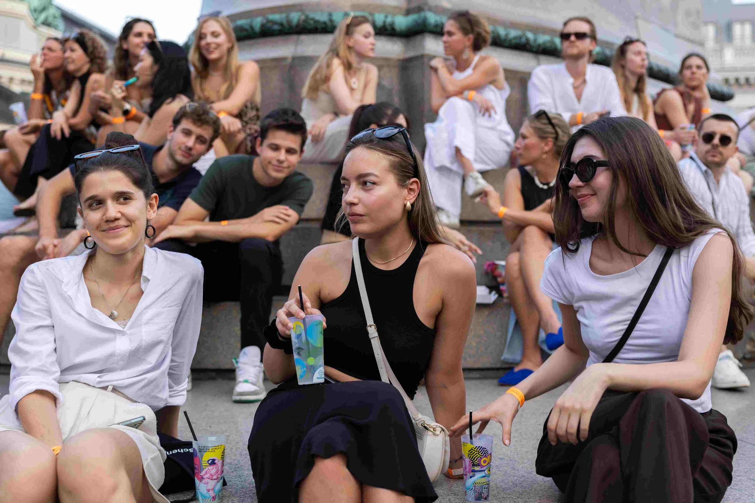 A group of young adults sitting on steps outdoors, some smiling and chatting, some wearing sunglasses, with a historic building in the background.