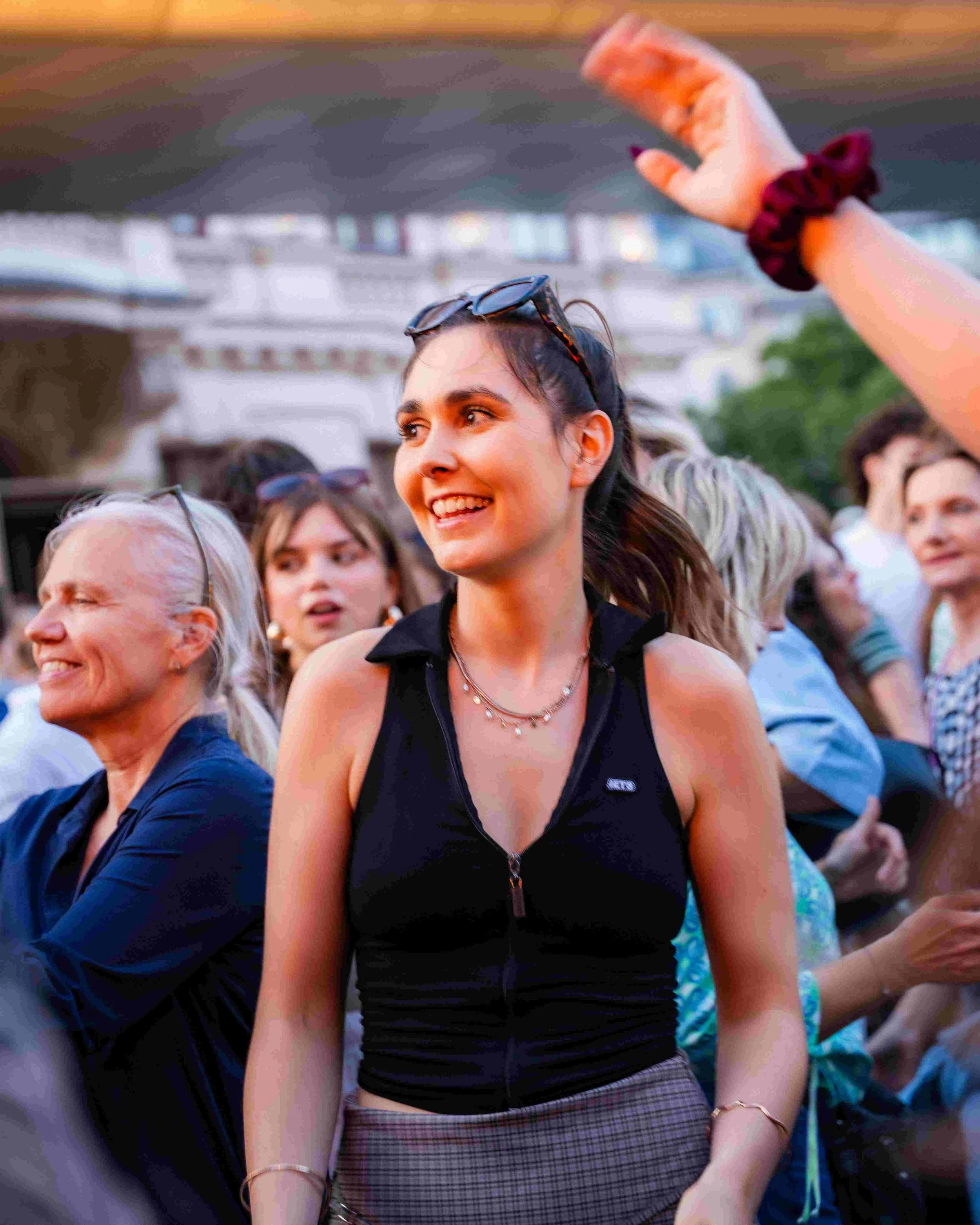 A woman with dark hair and sunglasses on her head, smiling at an outdoor event, surrounded by a crowd of people.
