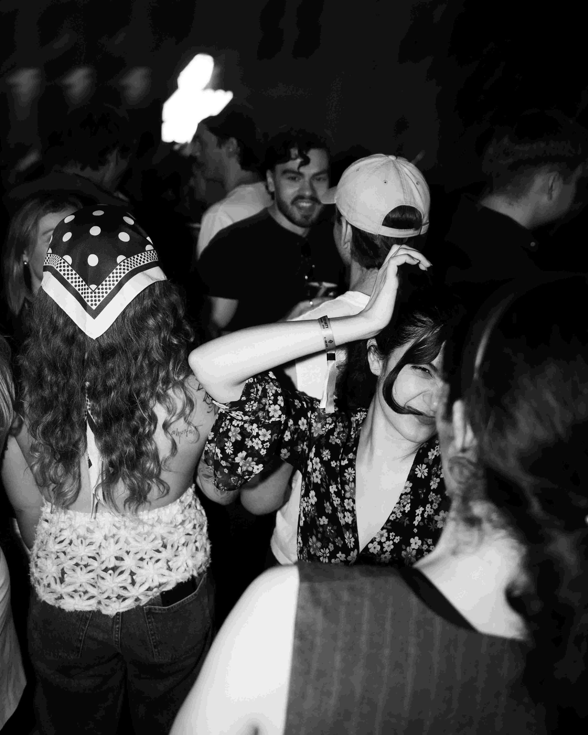 Group of young people at a party or social gathering, some dancing and talking, with one girl wearing a bandana, another girl with curly hair and a floral top, and a guy wearing a baseball cap.