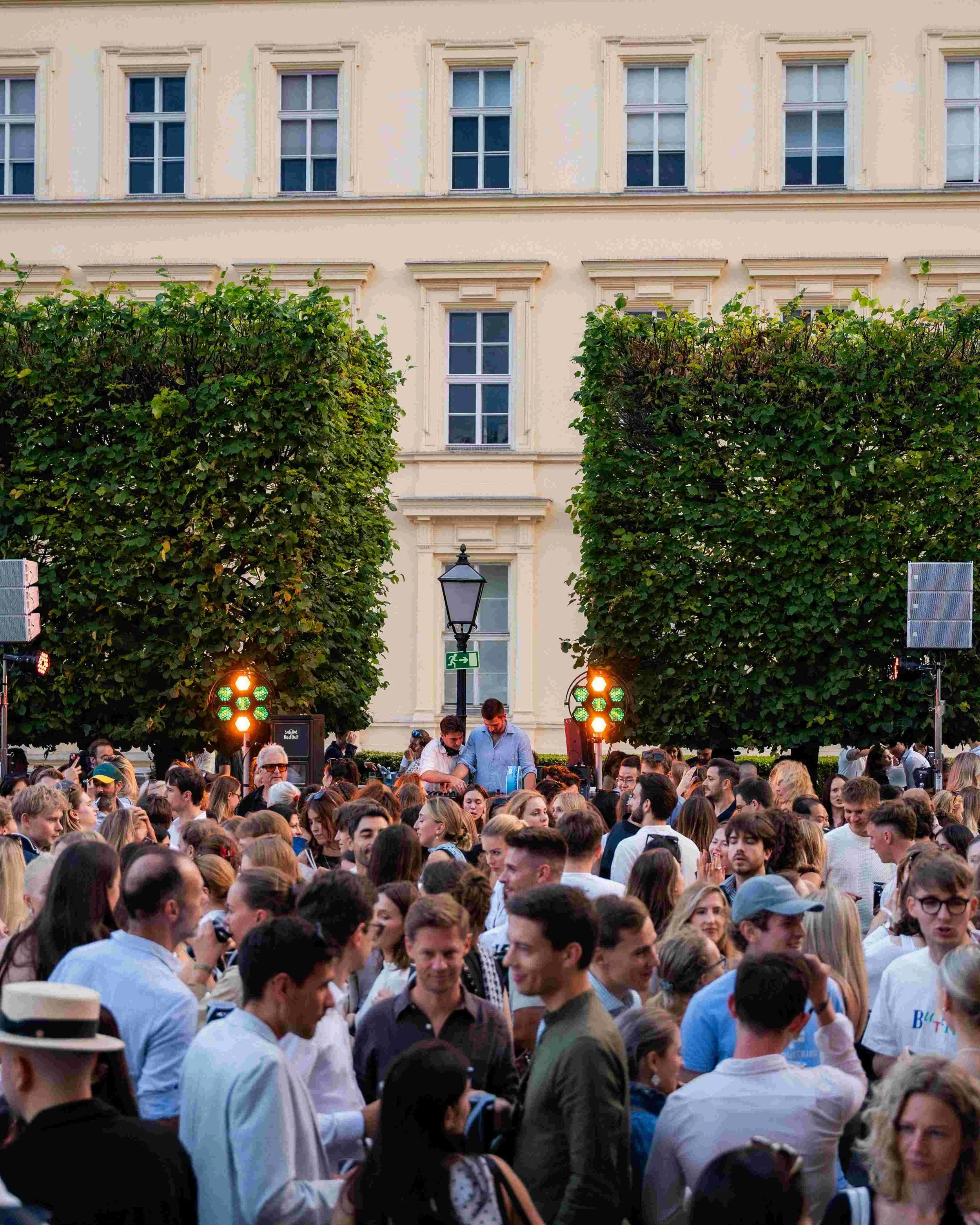 Crowd of people gathered outdoors in front of a building with multiple top-floor windows, green trees, street lamps, and stage lights.
