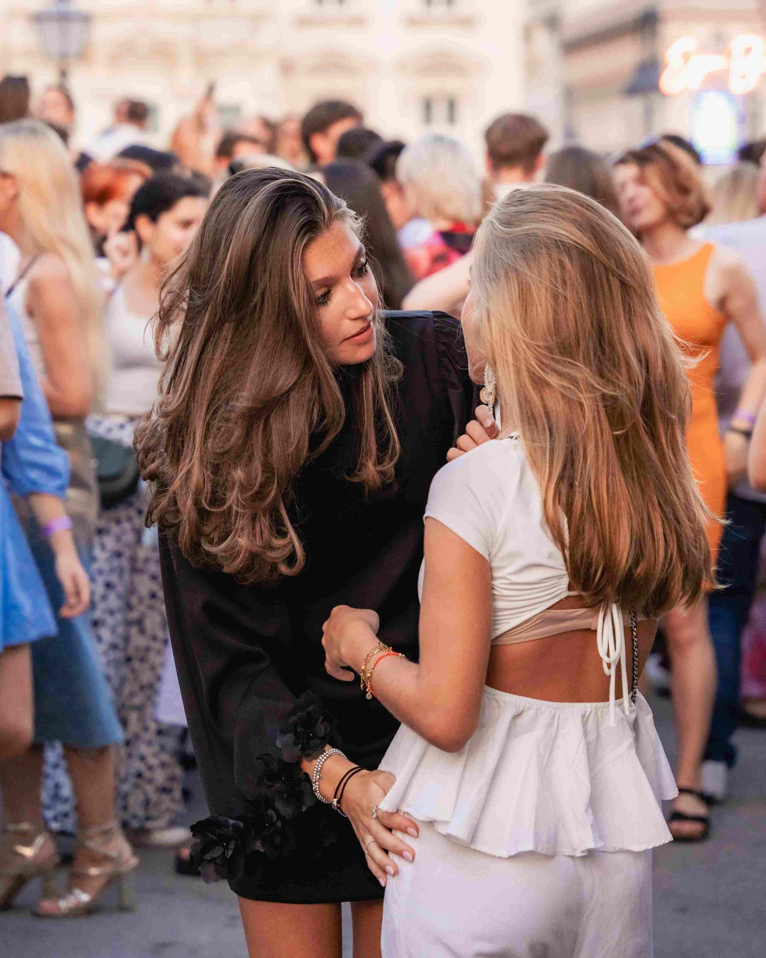 Two women are dancing and close to each other at a crowded indoor event. The woman on the left has long brown hair and is wearing a black dress. The woman on the right has long red hair and is wearing a white crop top and skirt. People are gathered i
