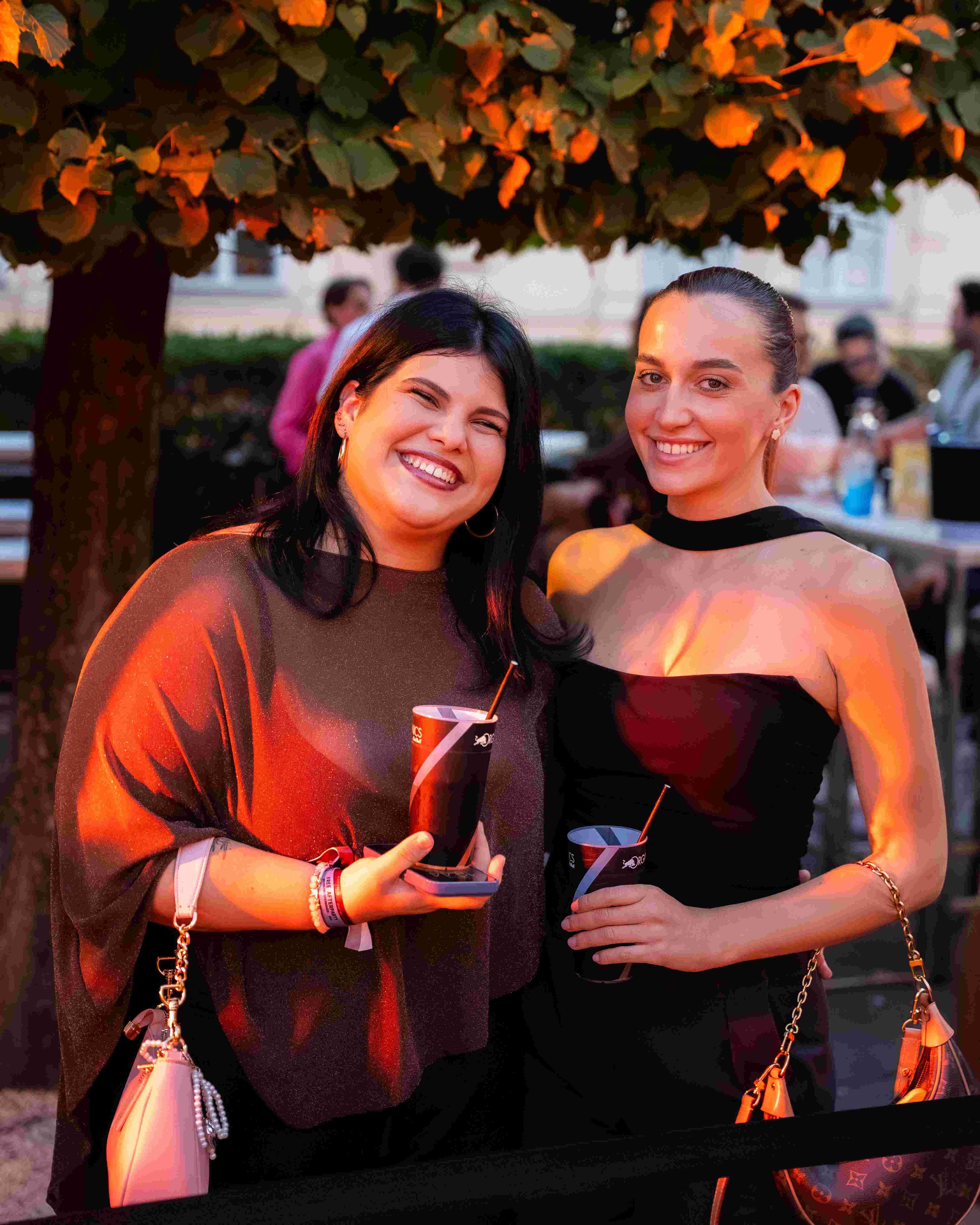 Two smiling women at an outdoor gathering holding drinks, under a tree with orange leaves.