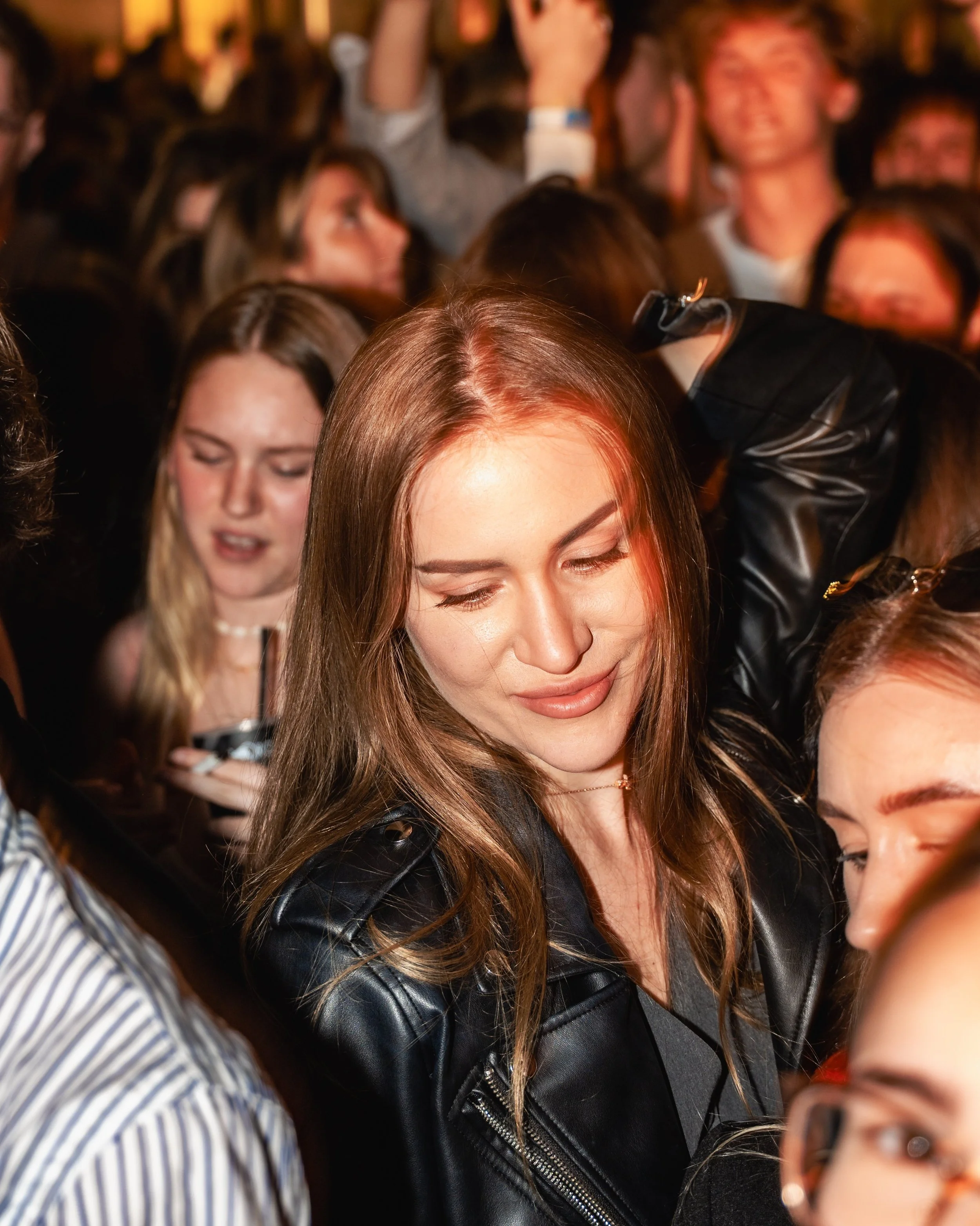 Close-up view of a young woman with long brown hair, wearing a black leather jacket, with other people in the background at what appears to be a crowded indoor event.