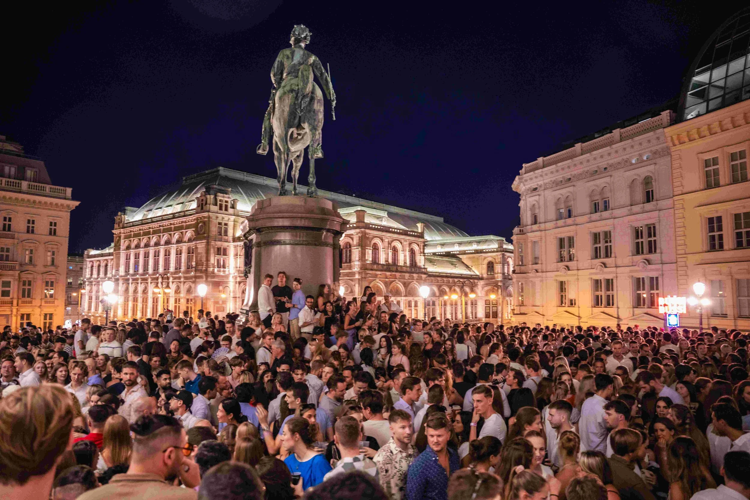 A large crowd of people gathered at night in a city square with historic buildings and a statue on horseback.