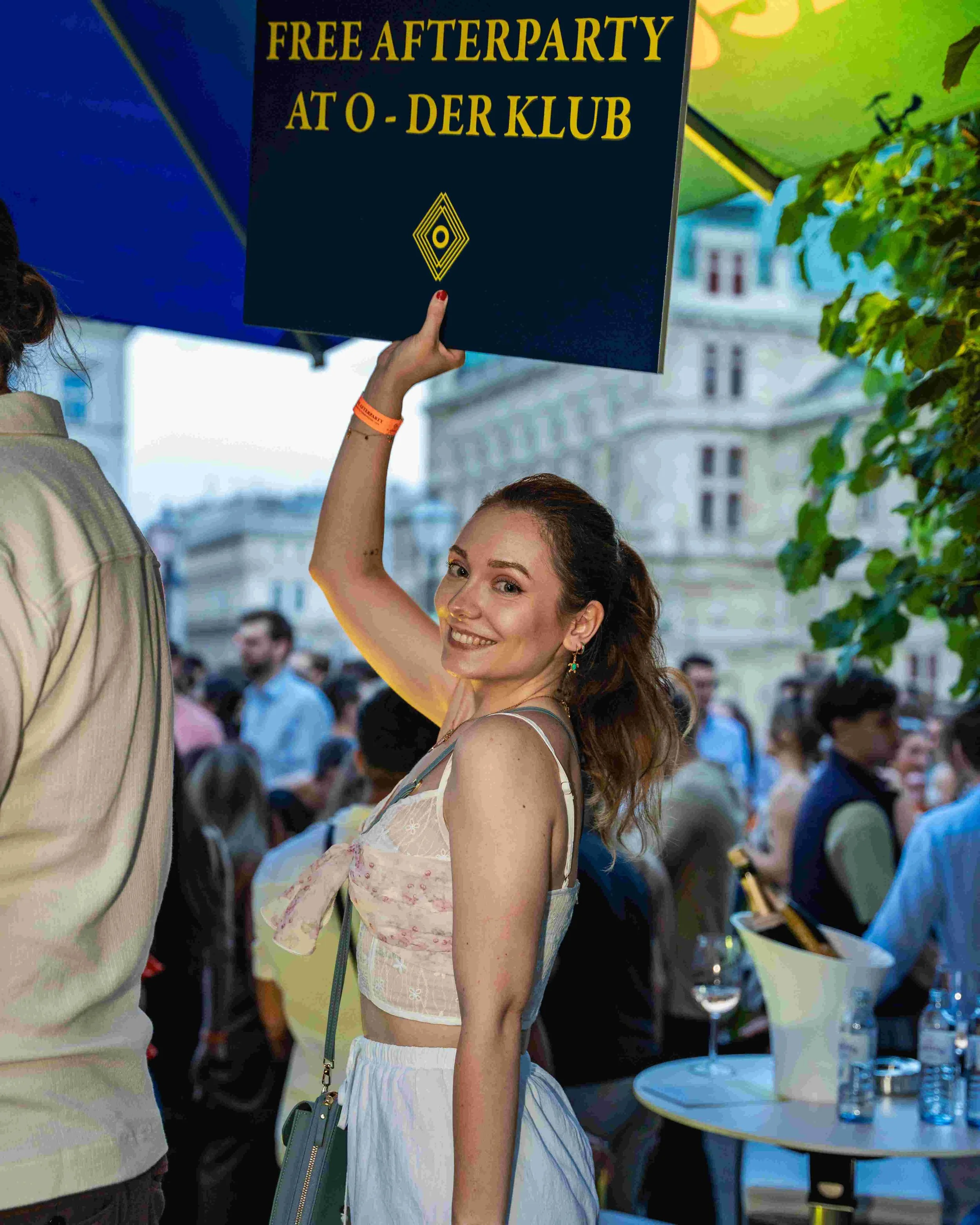 A young woman with red hair smiling and looking at the camera, holding a sign that reads "FREE AFTERPARTY AT O - DER KLUB" at an outdoor event during daytime, with other people and buildings in the background.