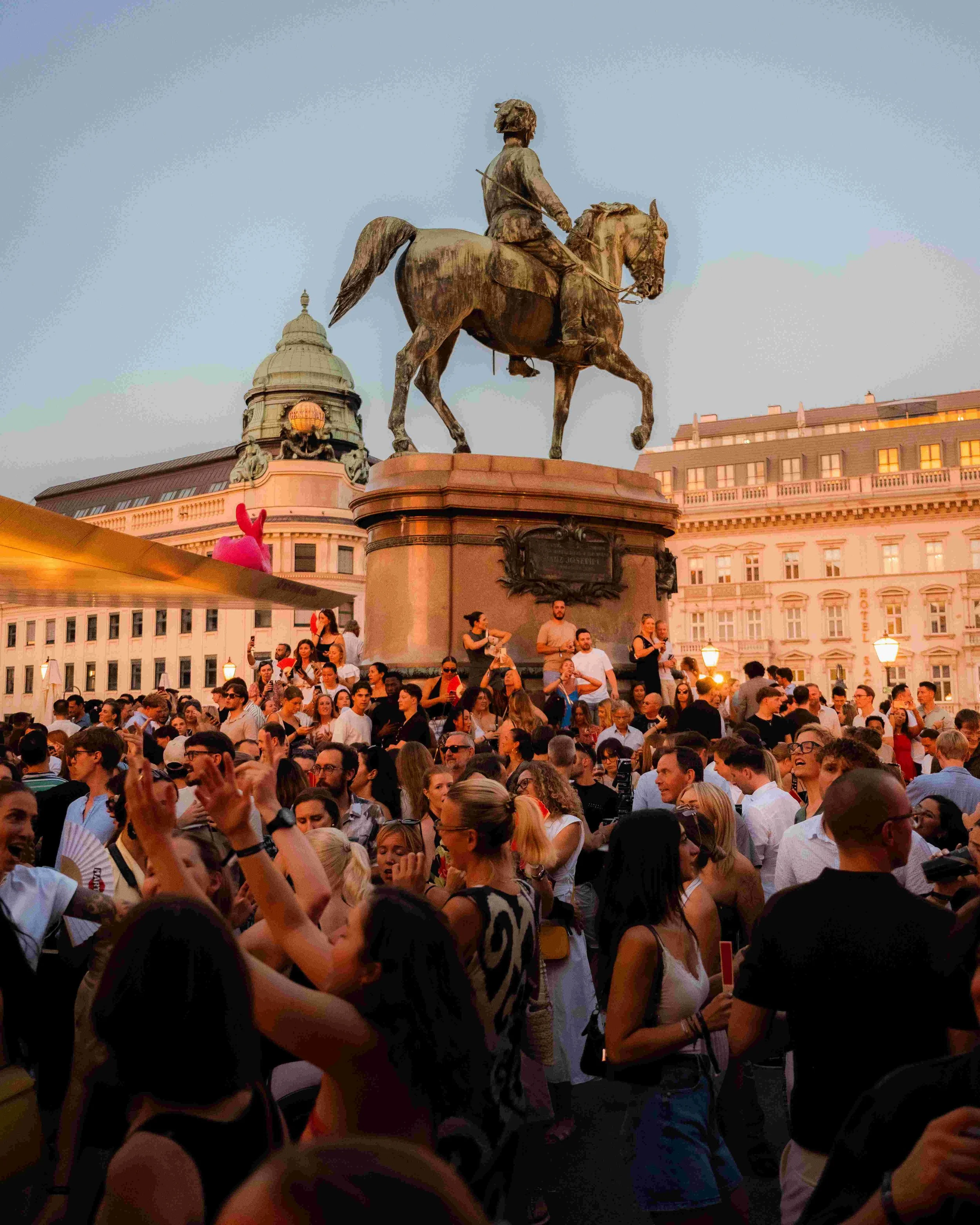 A large crowd of people gathered in a city square at dusk, with a prominent statue of a man on a horse on a pedestal in the background. The crowd appears to be attending an event or celebration.