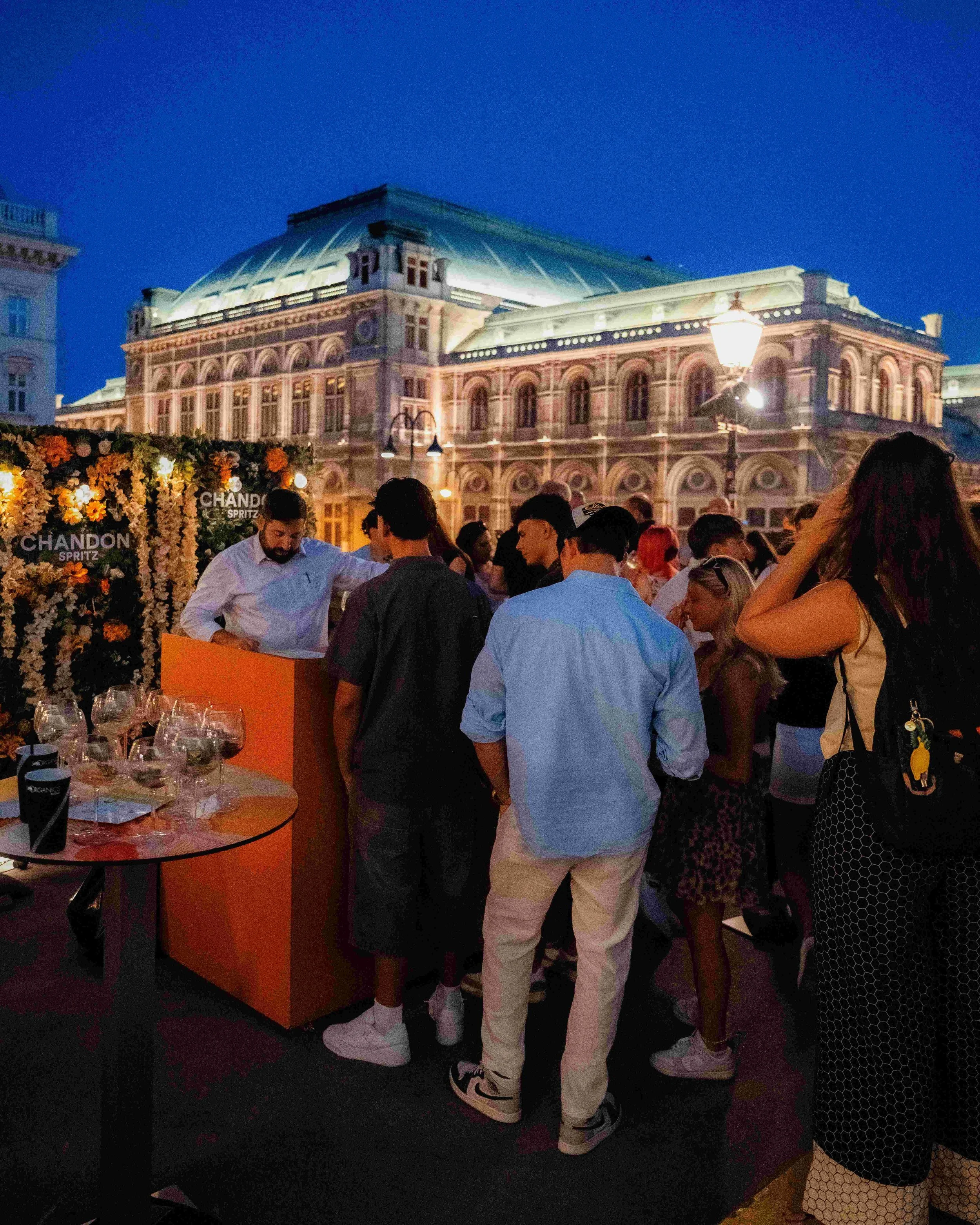 People gathered at an outdoor event during the evening, with a historic building illuminated in the background. There are tables with glasses and a floral decoration with a 'Chandon Spritz' sign in the foreground.