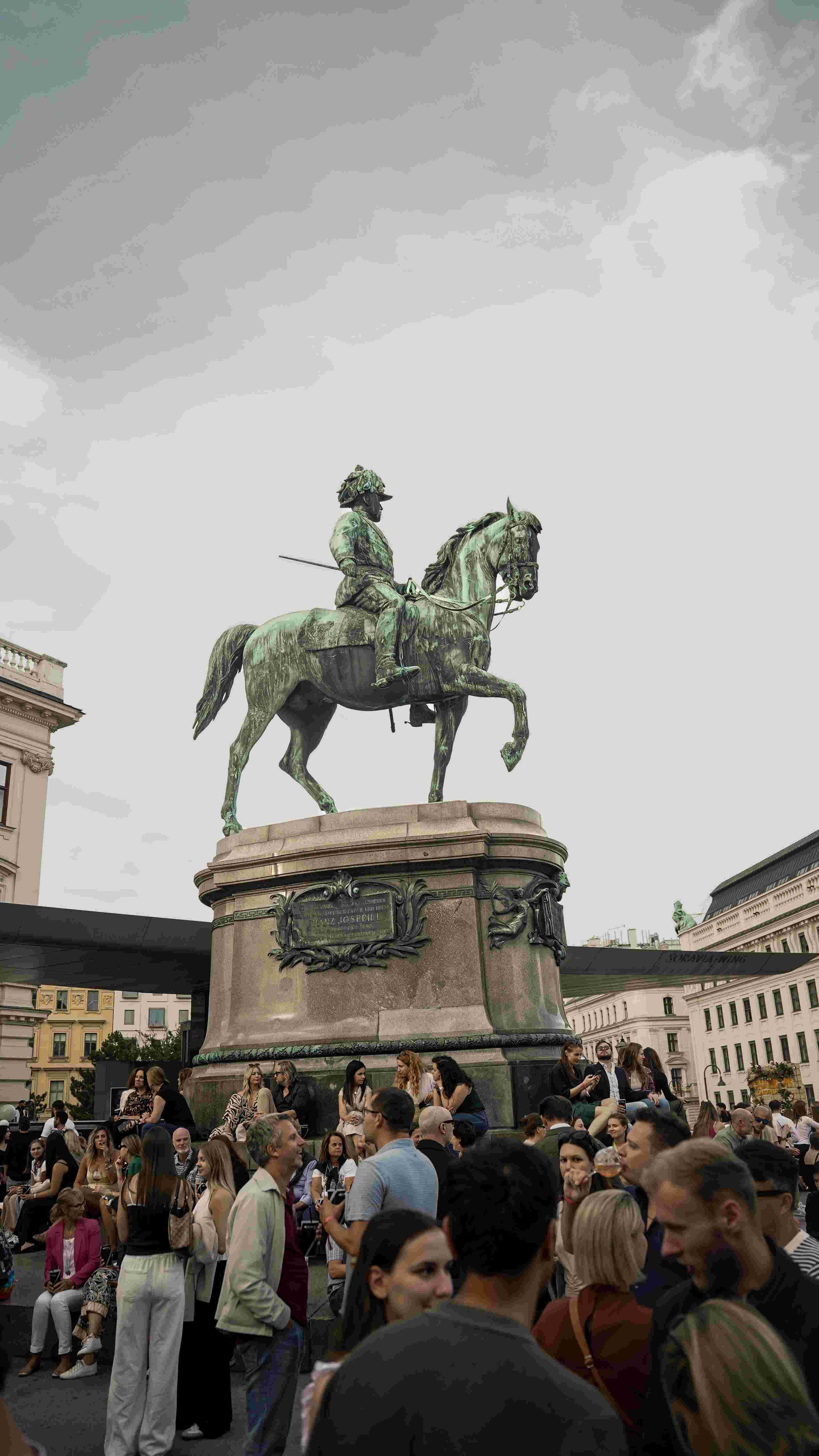 A crowded square with people gathered around an equestrian statue of a man on a horse, set against gray sky and surrounding buildings.