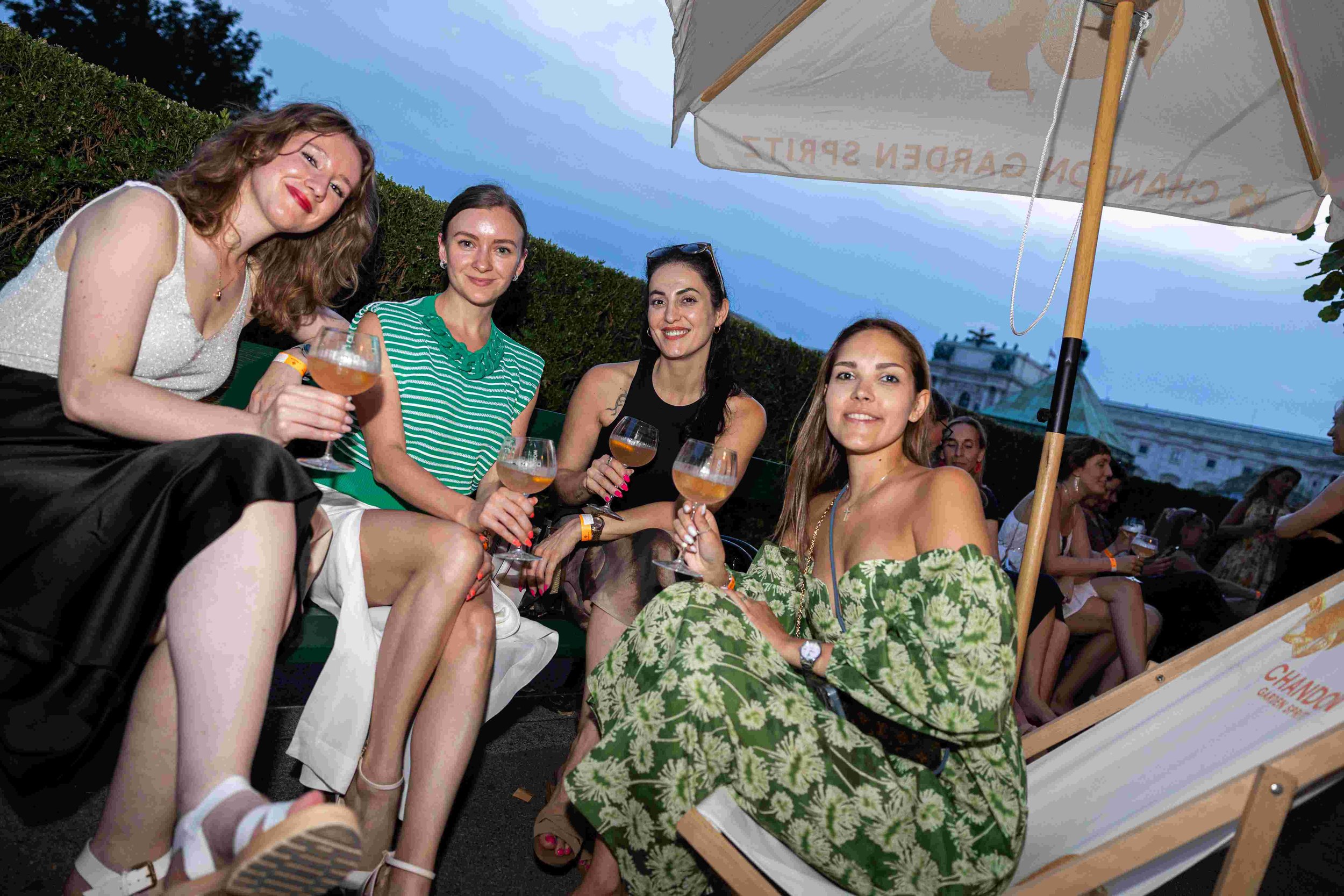 Four women sitting outdoors under a large white patio umbrella, holding drinks, smiling, in front of a hedge and a building, during evening.