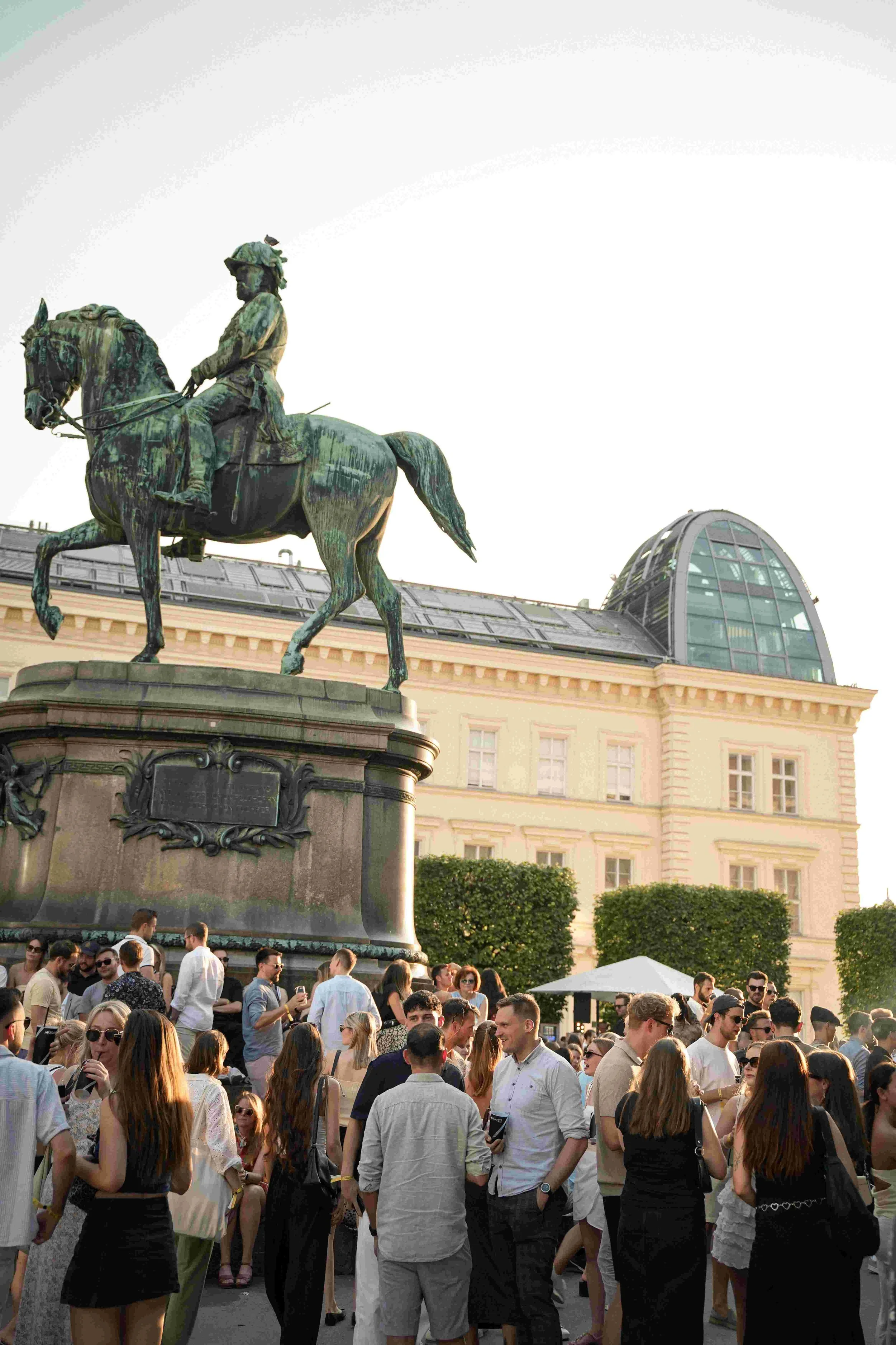 Crowd of people gathered outdoors around a large equestrian statue of a historical figure on a horse, with a historic building and modern architecture in the background.
