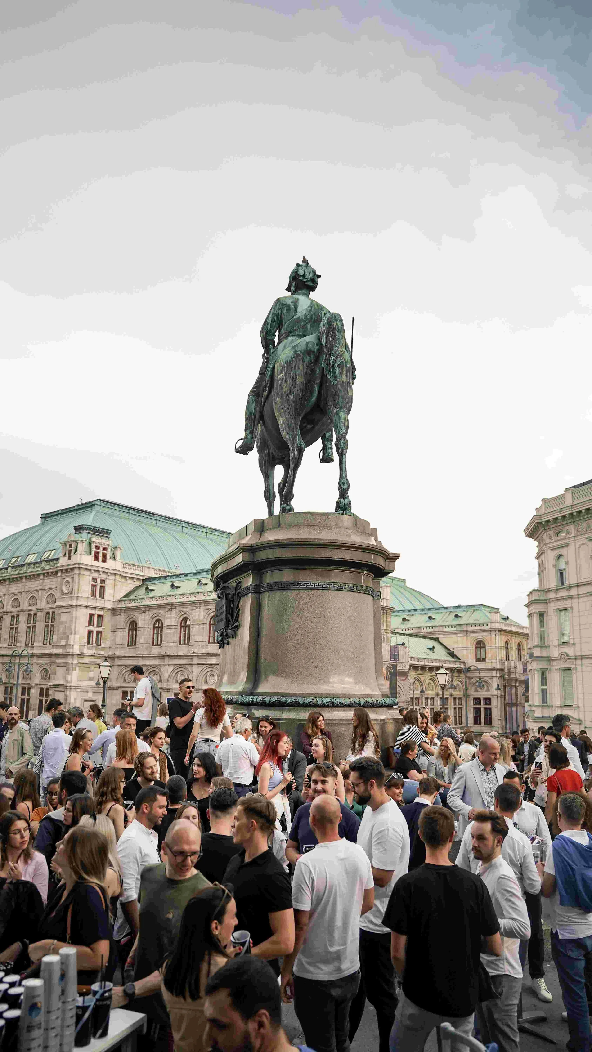 Crowd gathered around a large equestrian statue in an urban square with historic buildings and a cloudy sky in the background.