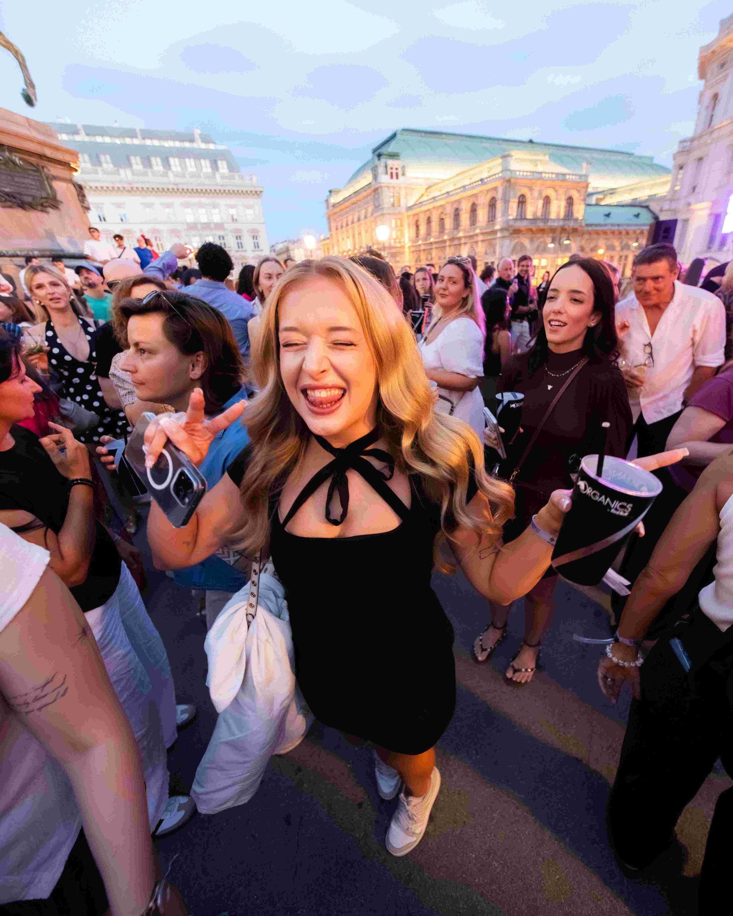 People gathered at an outdoor event in an urban setting during the evening. A smiling woman with blonde hair in the foreground is making a playful face, holding a beverage and a phone. Other attendees are talking and socializing, with historic buildi