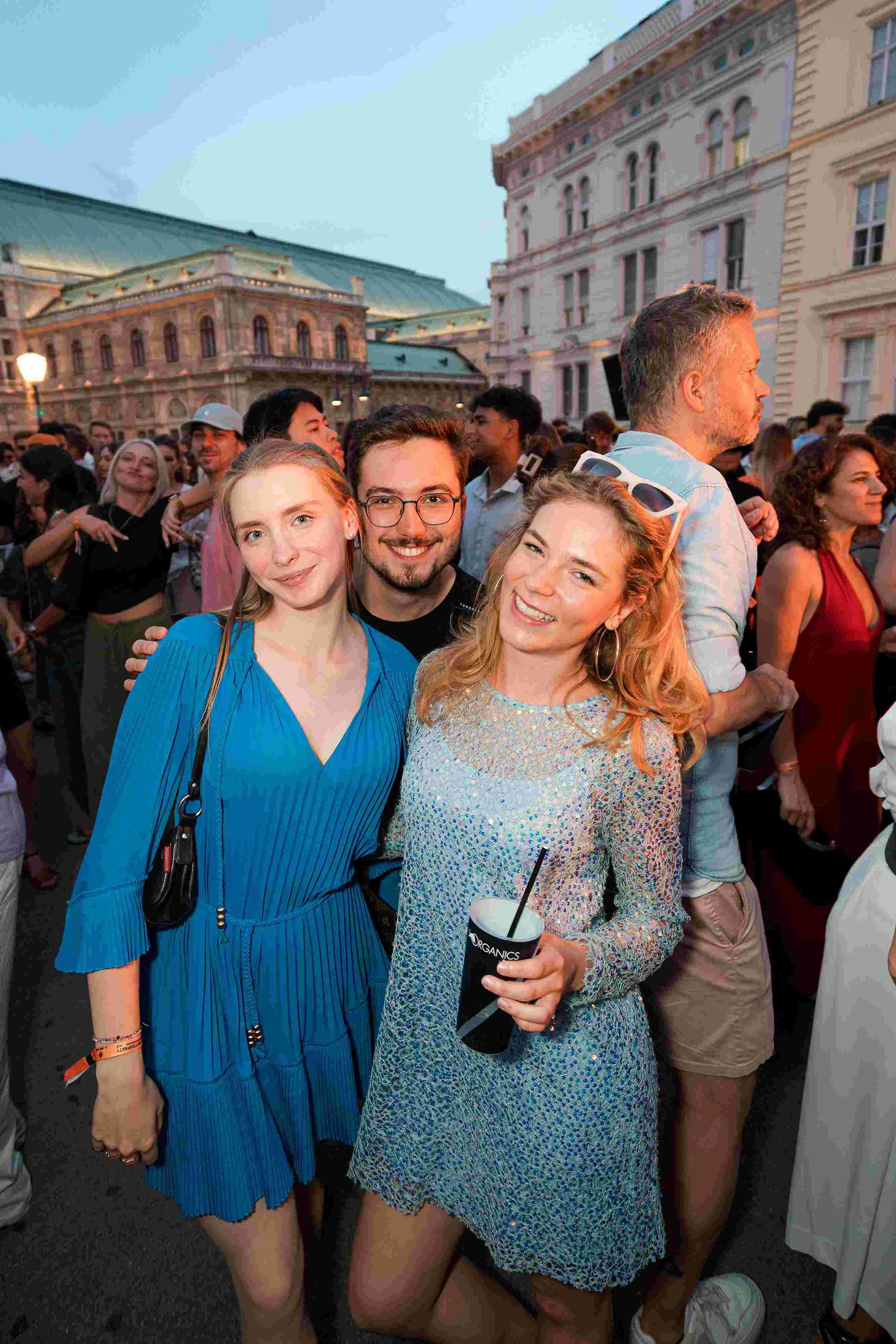 A group of young adults attending an outdoor event or festival in front of historical buildings during evening time. The focus is on three women and a man smiling at the camera, with many other people in the background.