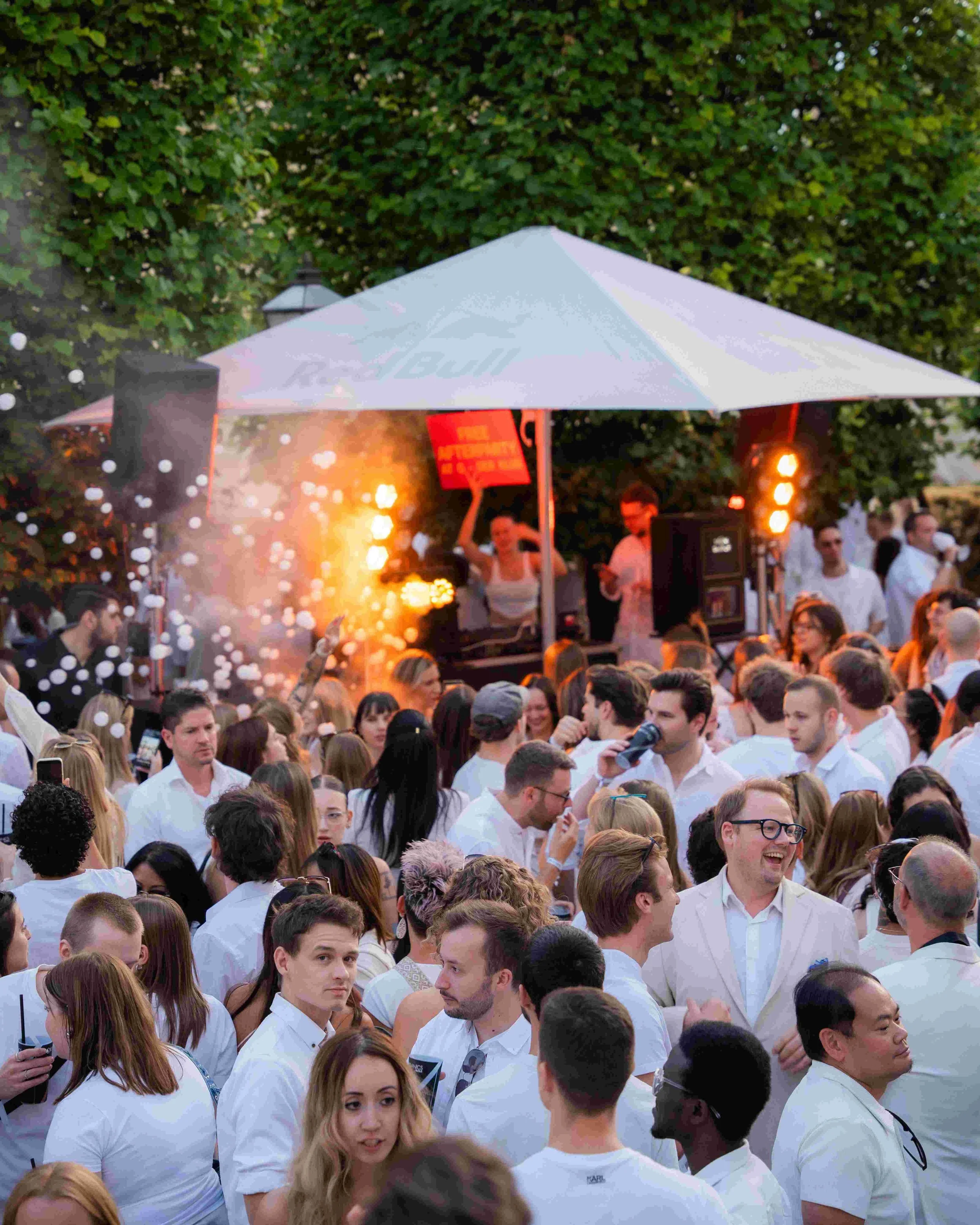 A crowd of people gathered outdoors during the evening, many dressed in white, with some smiling and talking, under a large white canopy with a sign that reads 'BULM'. There are green trees in the background and stage lights creating an energetic atm