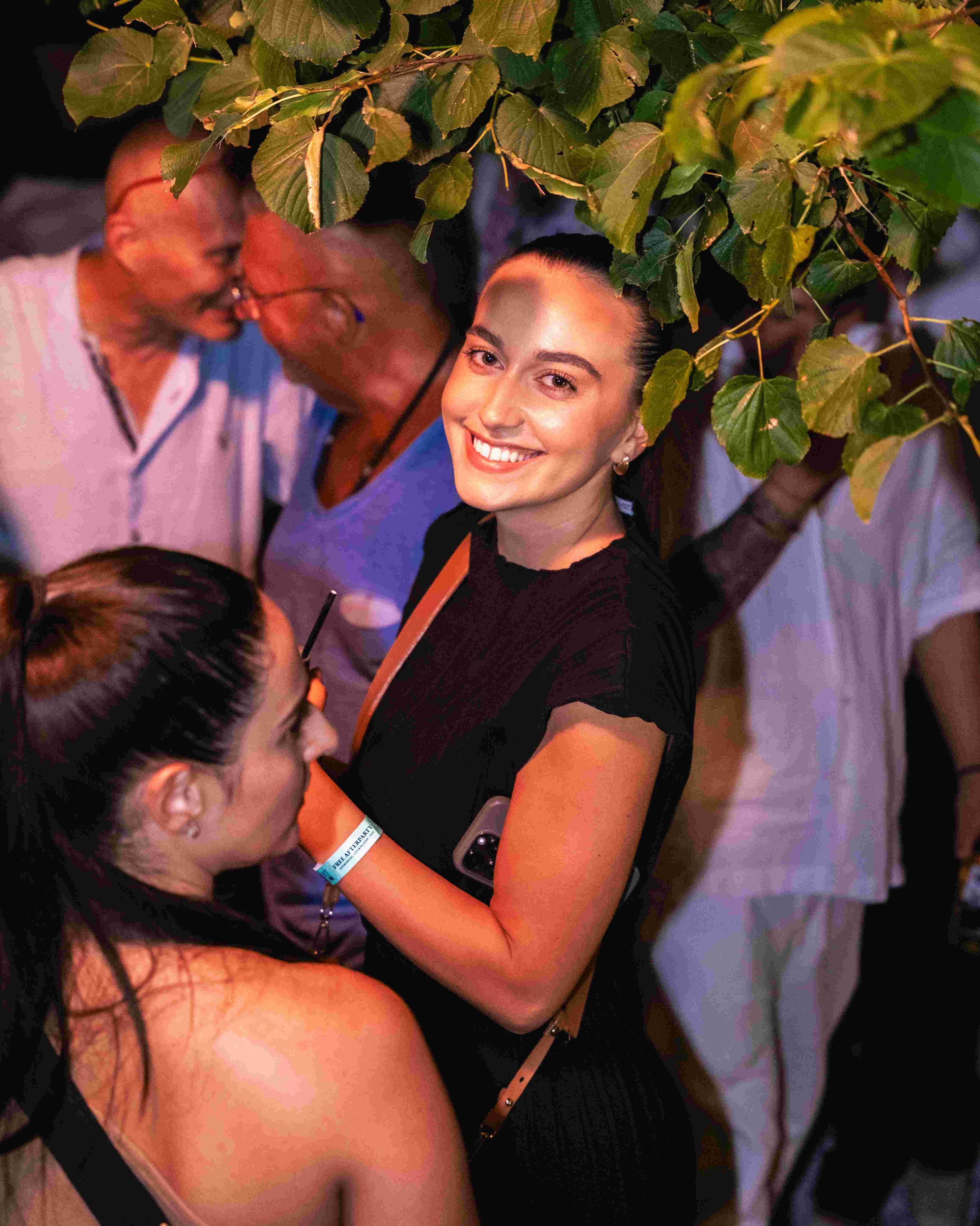 A group of people at a social gathering, with a young woman smiling and taking a selfie while surrounded by others, some wearing glasses and colorful clothing, under a tree with green leaves.