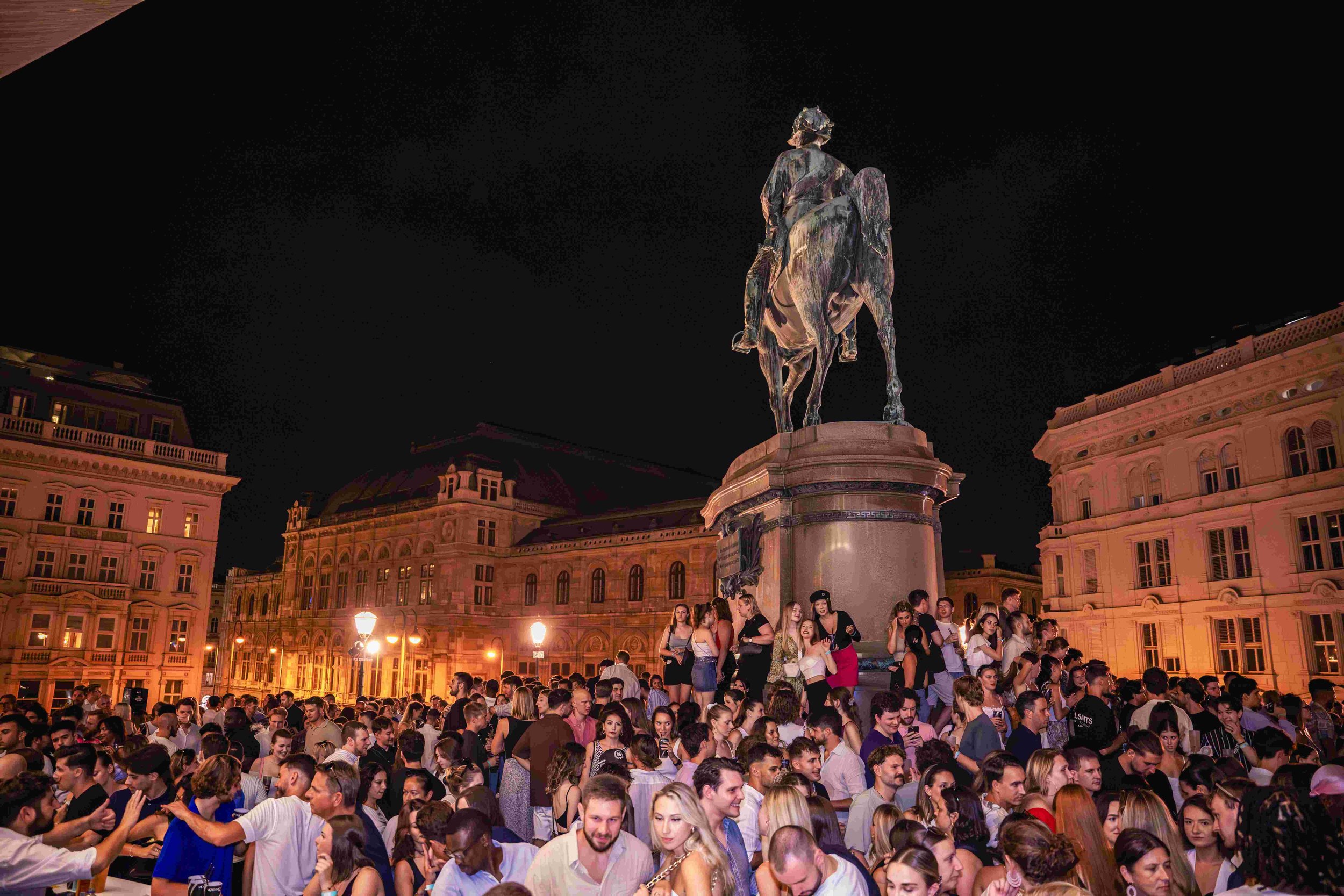 Nighttime scene in a city square with a large crowd of people gathered, standing and socializing, in front of an equestrian statue on a tall pedestal surrounded by historic buildings with lit windows.