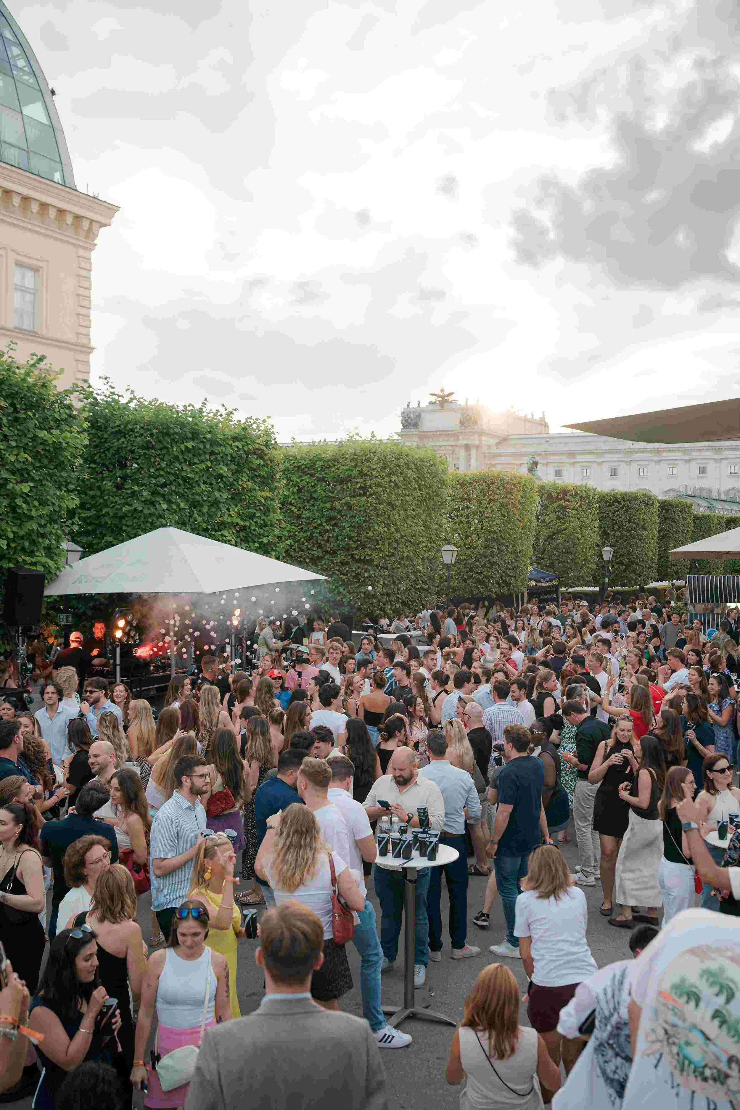 A large crowd of people gathered outdoors in front of historic buildings with green trees and cloudy sky, at what appears to be a lively social event or concert.