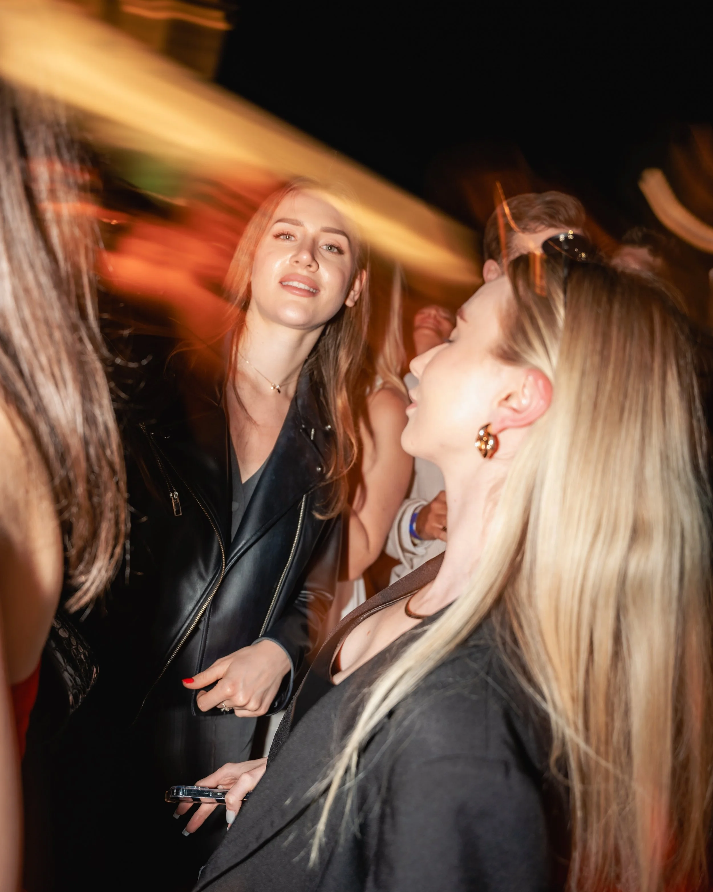 Group of women in a crowded setting at night with motion blur, some with glasses, engaged in conversation.