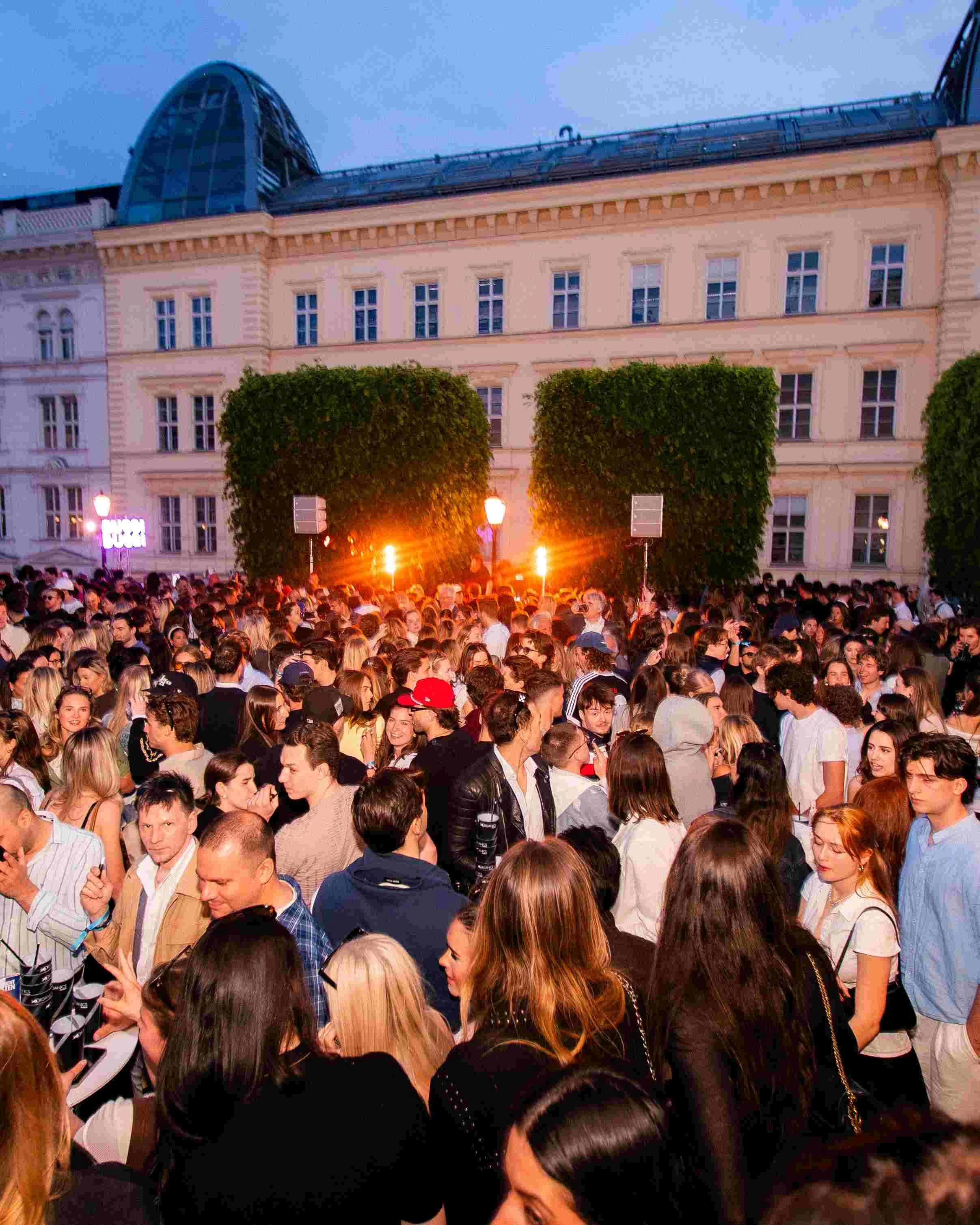 A large crowd of people gathered outdoors during the evening, with some engaging with mobile devices, and a building with multiple windows and a distinctive glass dome on top in the background. There are trees and street lamps illuminating the scene,
