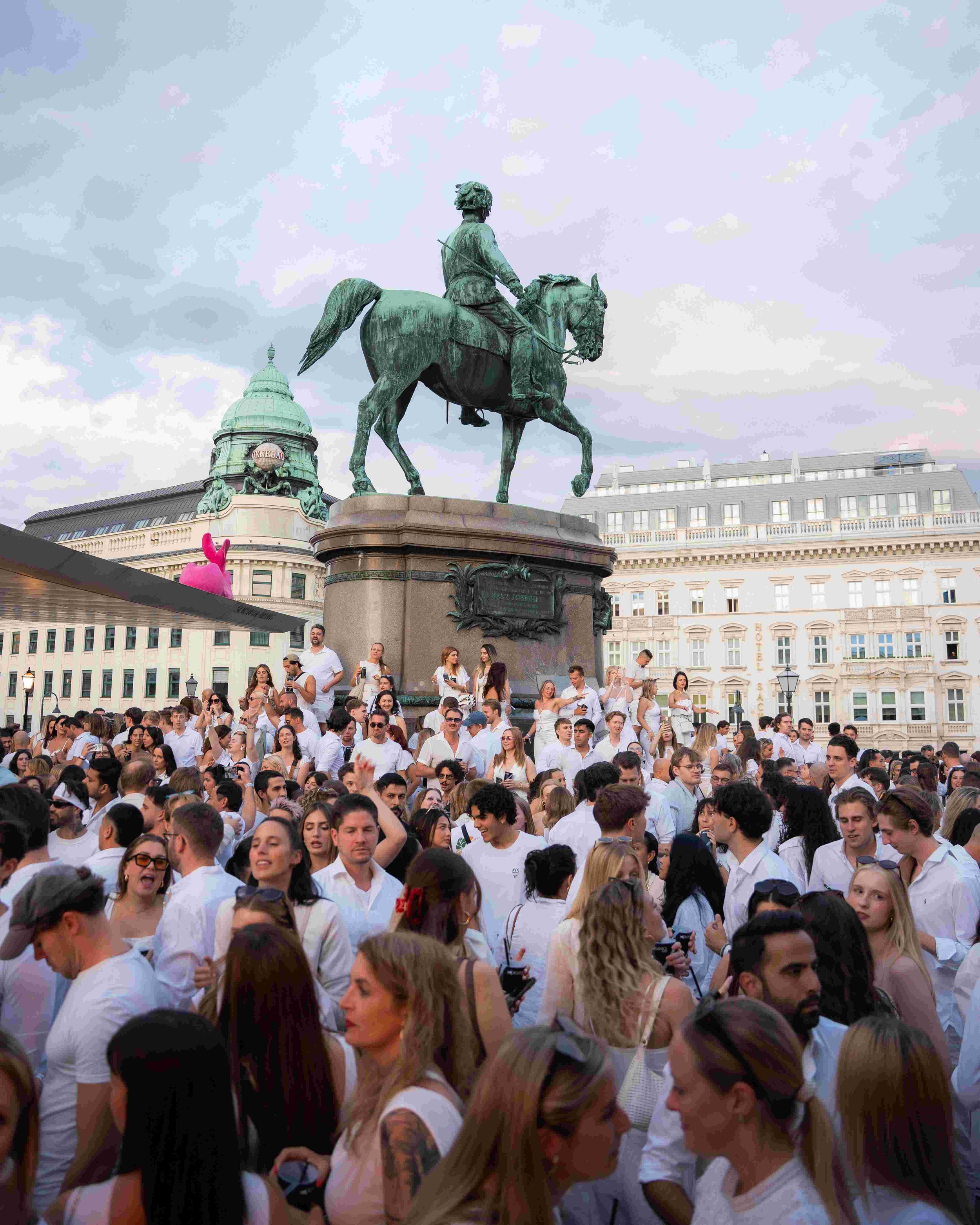 A large crowd of people gathered in an outdoor public square, wearing white clothing. A prominent equestrian statue of a man on horseback is in the background, with historic buildings and a cloudy sky above.