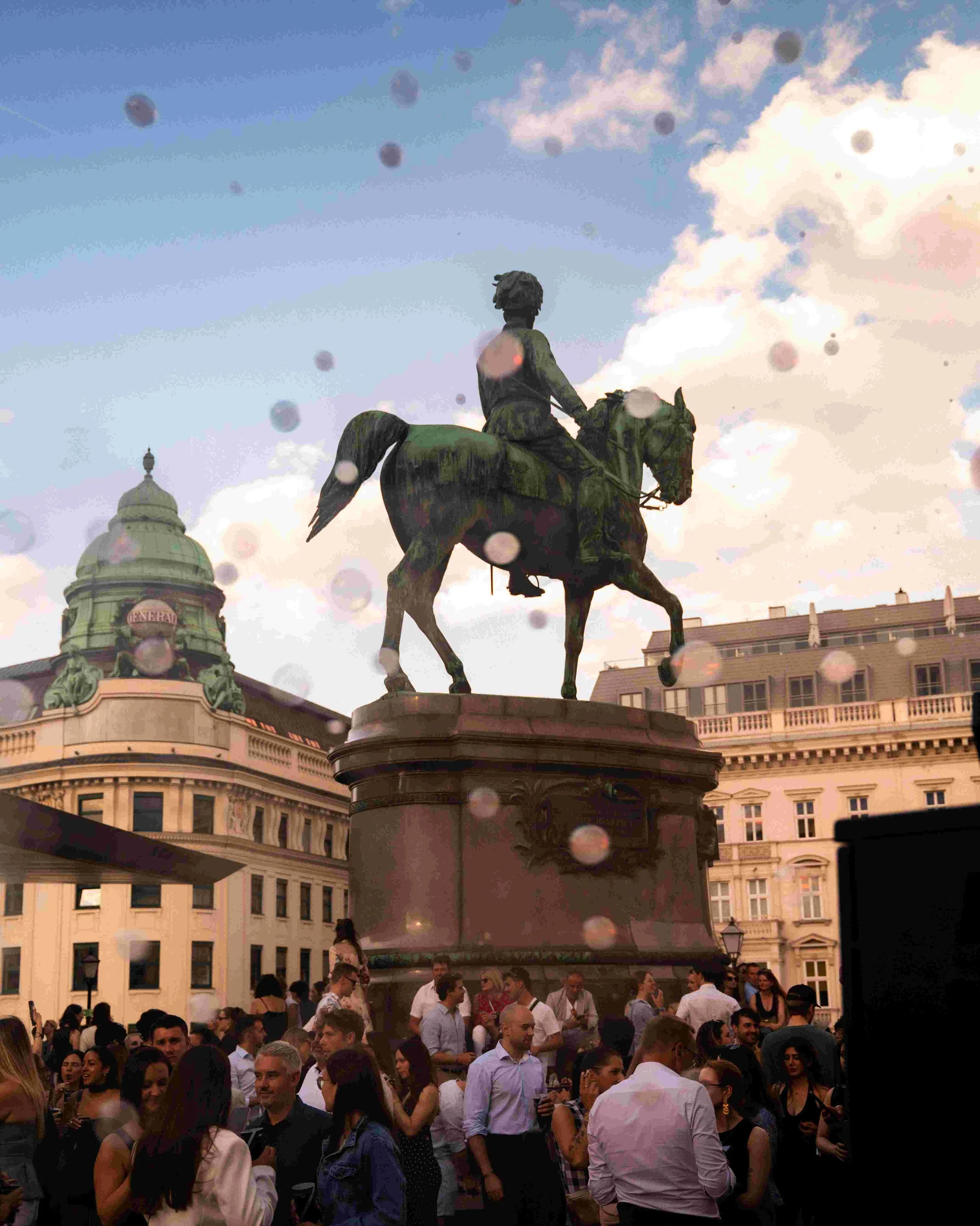 Crowd of people gathered around an equestrian statue in an urban square during daytime, with a building and a dome with the word 'GENERALI' in the background, and a partly cloudy sky.