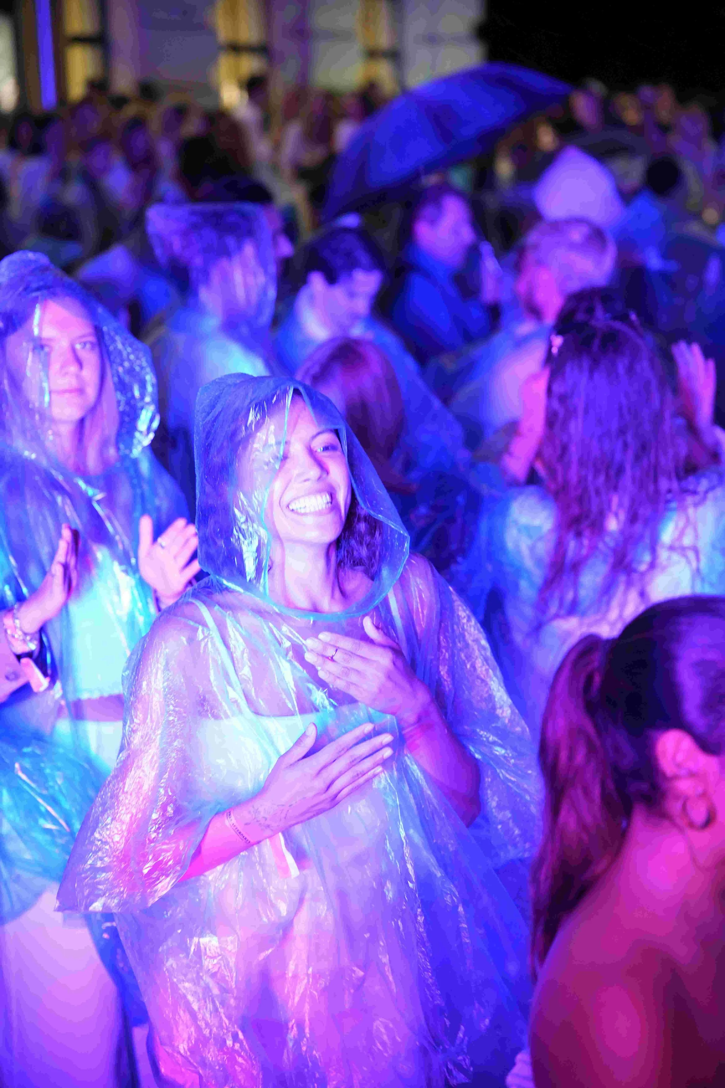 A group of people, mostly women, wearing clear plastic rain ponchos, stands close together in an indoor setting illuminated with purple and blue lights. Some have their hands over their hearts, and one woman is smiling with her eyes closed, appearing