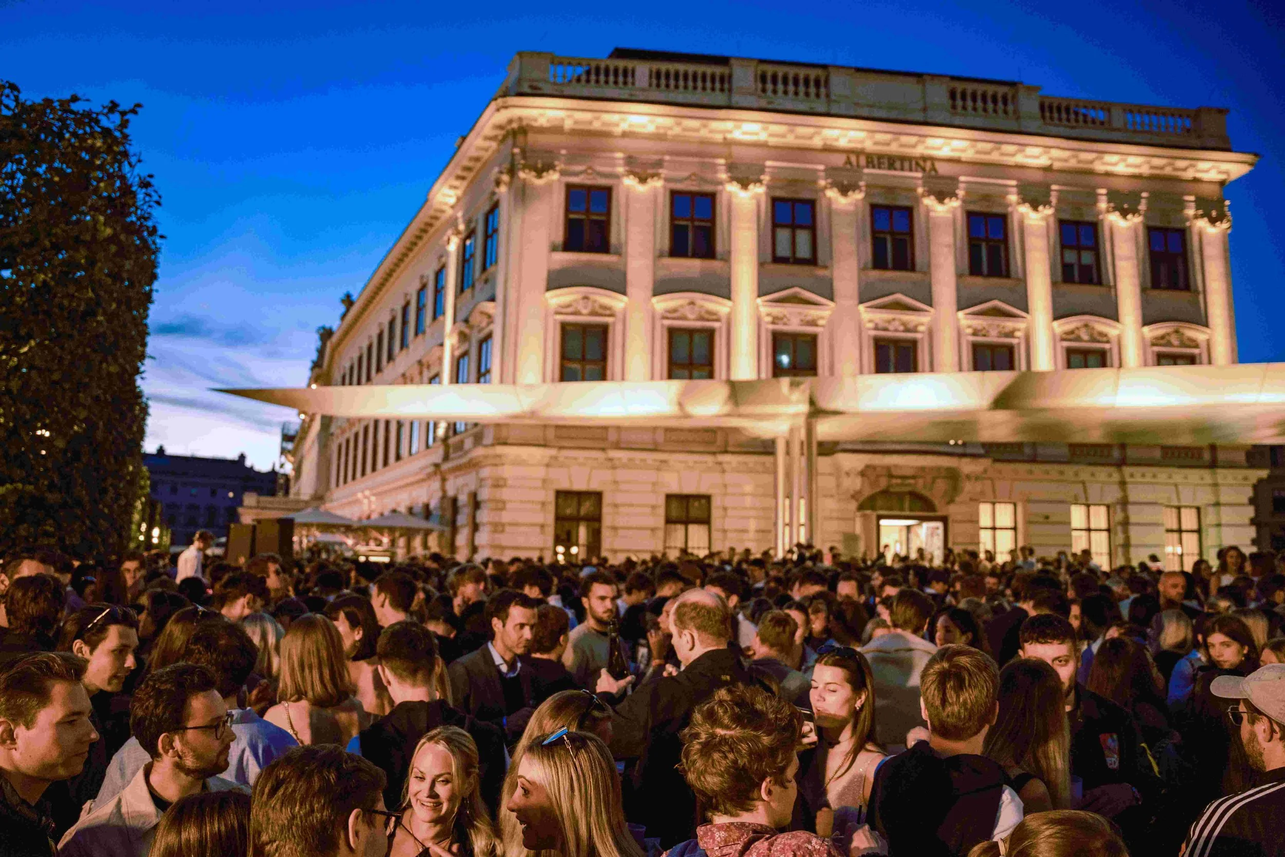 Crowd of people gathered outside a large, illuminated historic building during evening, with blue sky in background.