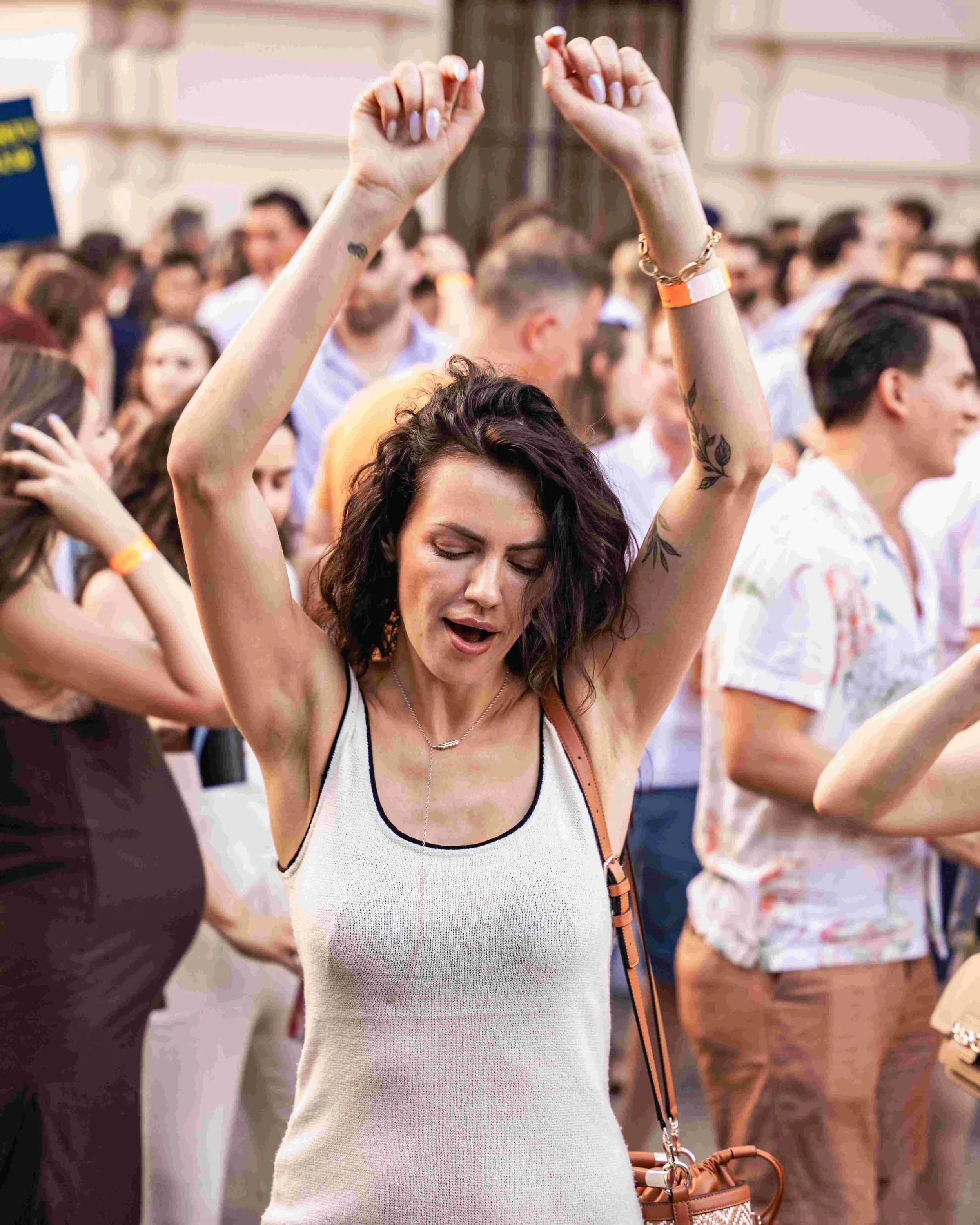 A woman with dark wavy hair and tattoos on her arms is dancing with her eyes closed at a crowded indoor event, raising her arms above her head in a joyful expression.