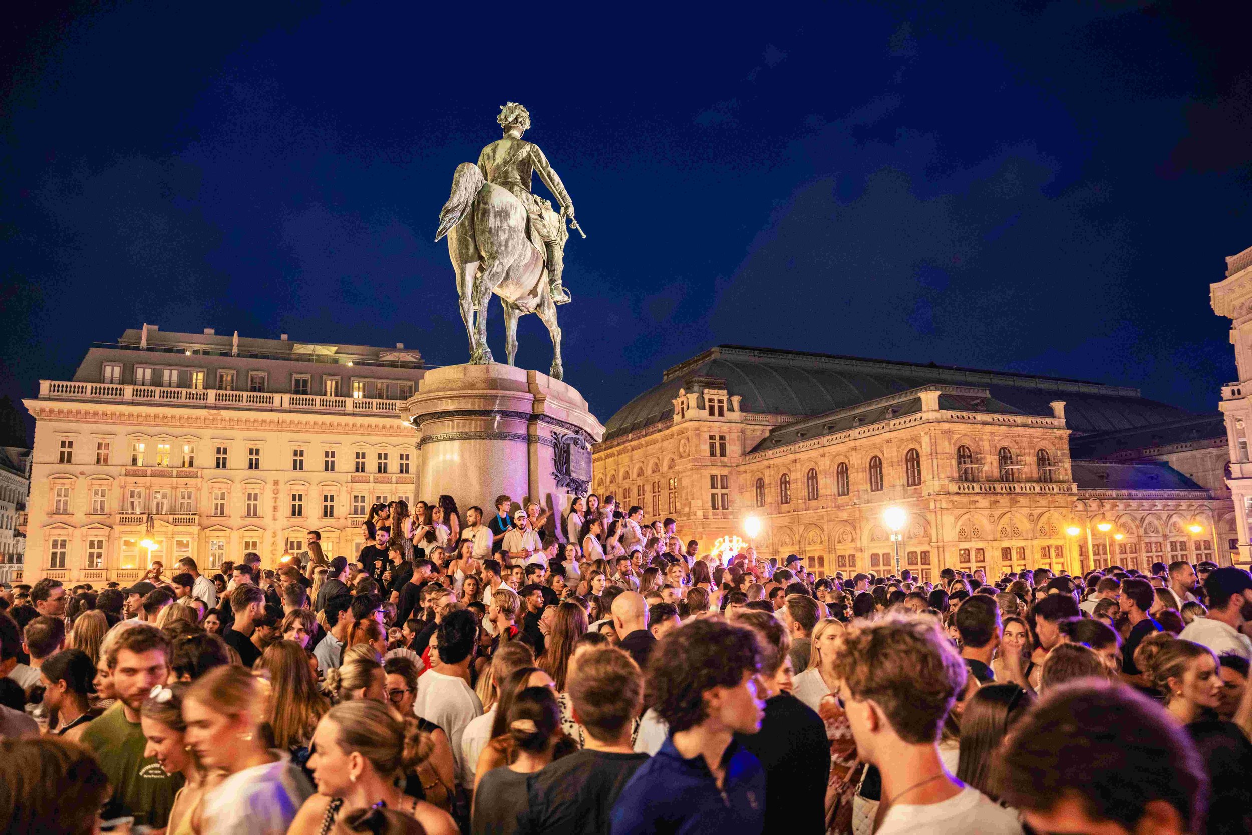 A large crowd of people gathered in a public square at night, with a statue of a man on horseback visible in the background and historic buildings surrounding the square.