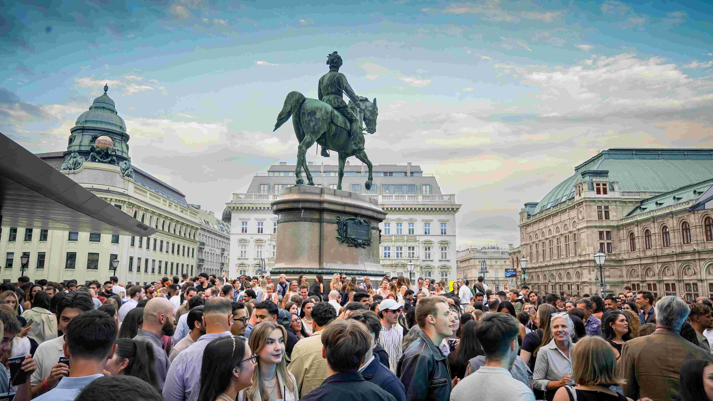 Crowds of people gathering in a city square under a cloudy sky, with a large equestrian statue and historic buildings in the background.