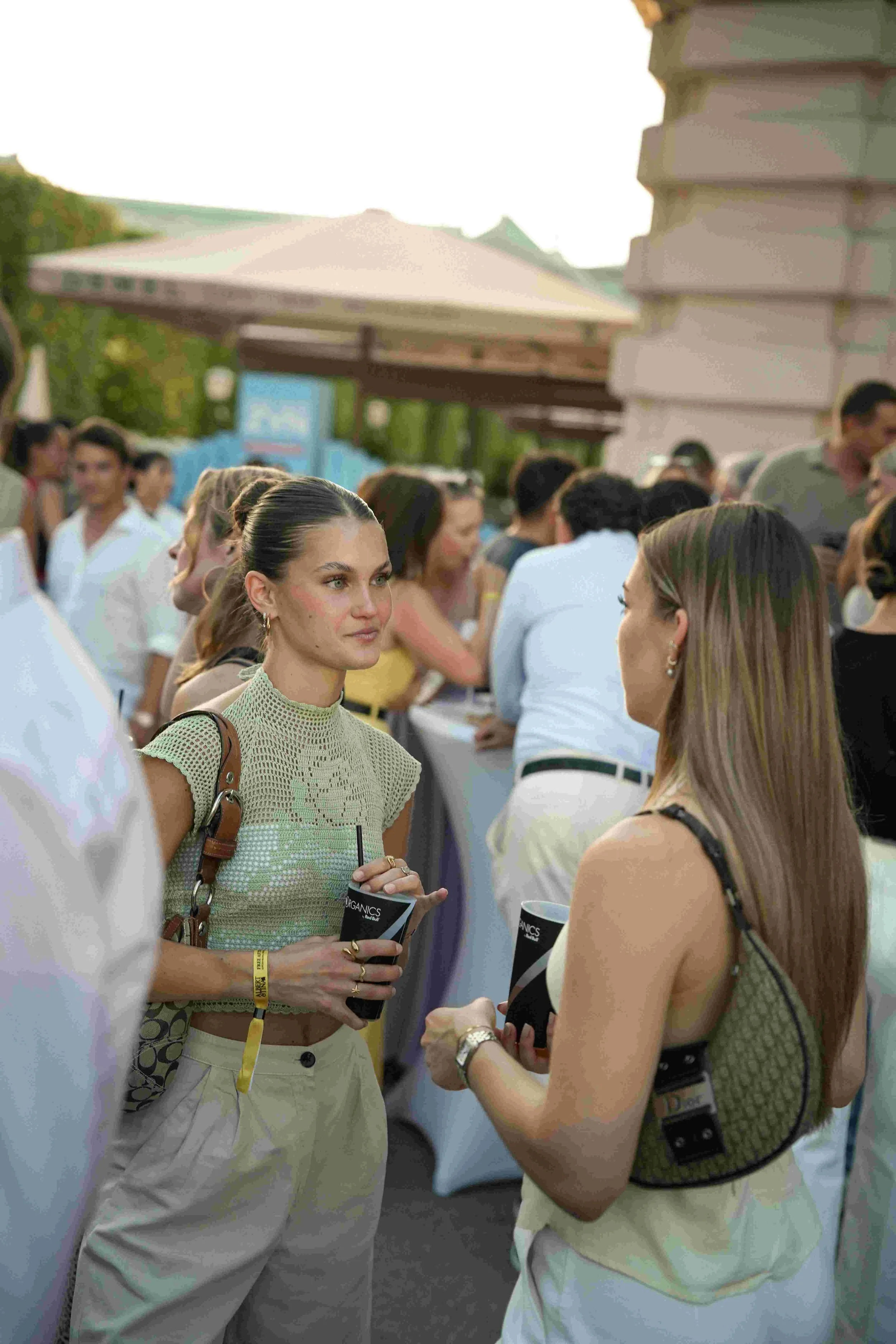 A group of people gathered outdoors at a social event, engaging in conversations. The focus is on a woman with brown hair, wearing a light green crocheted top and beige pants, holding a drink and talking to another woman with long brown hair. People 