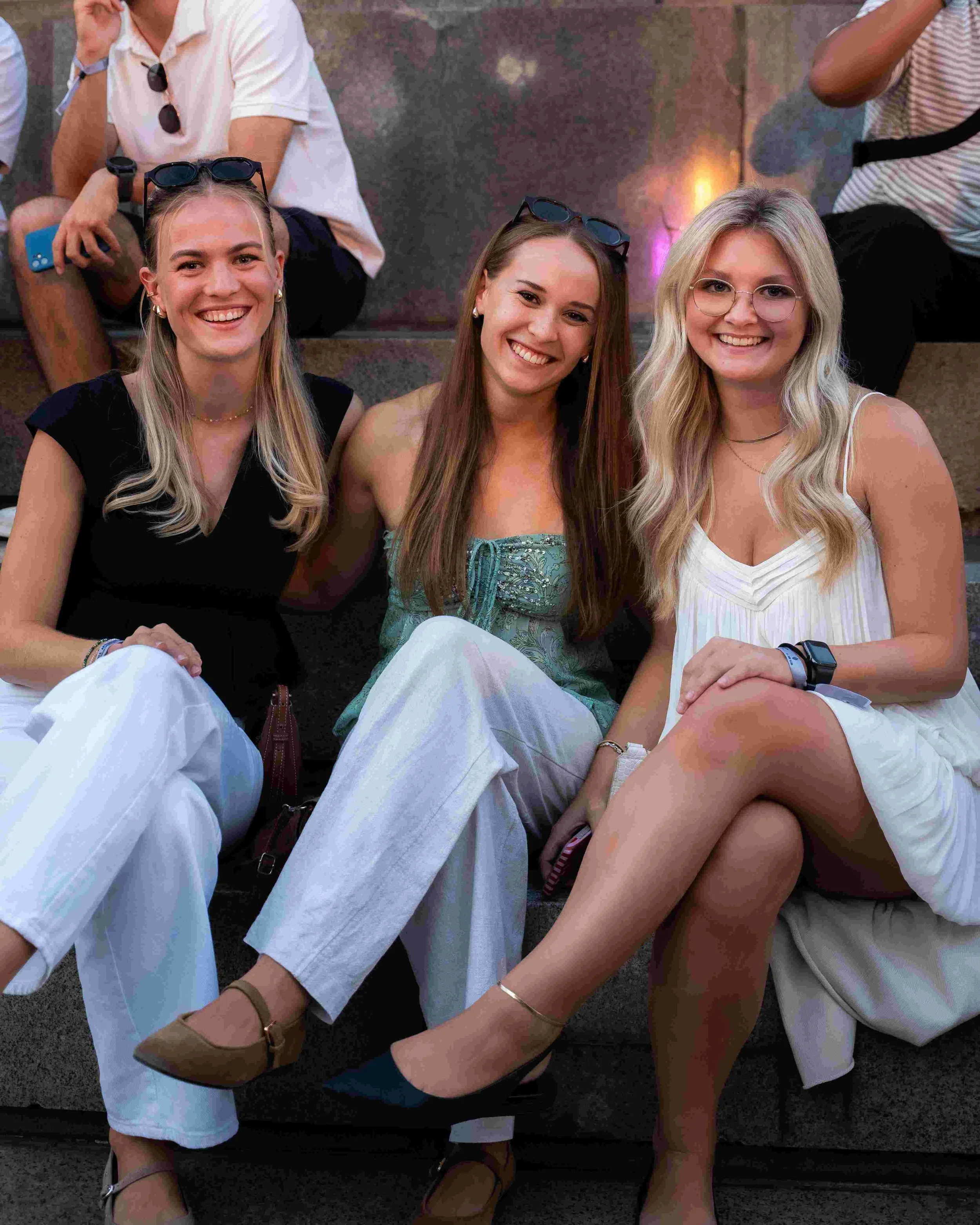 Three young women sitting together, smiling for the camera at what appears to be an outdoor gathering or event, with two wearing glasses and casual summer clothes.