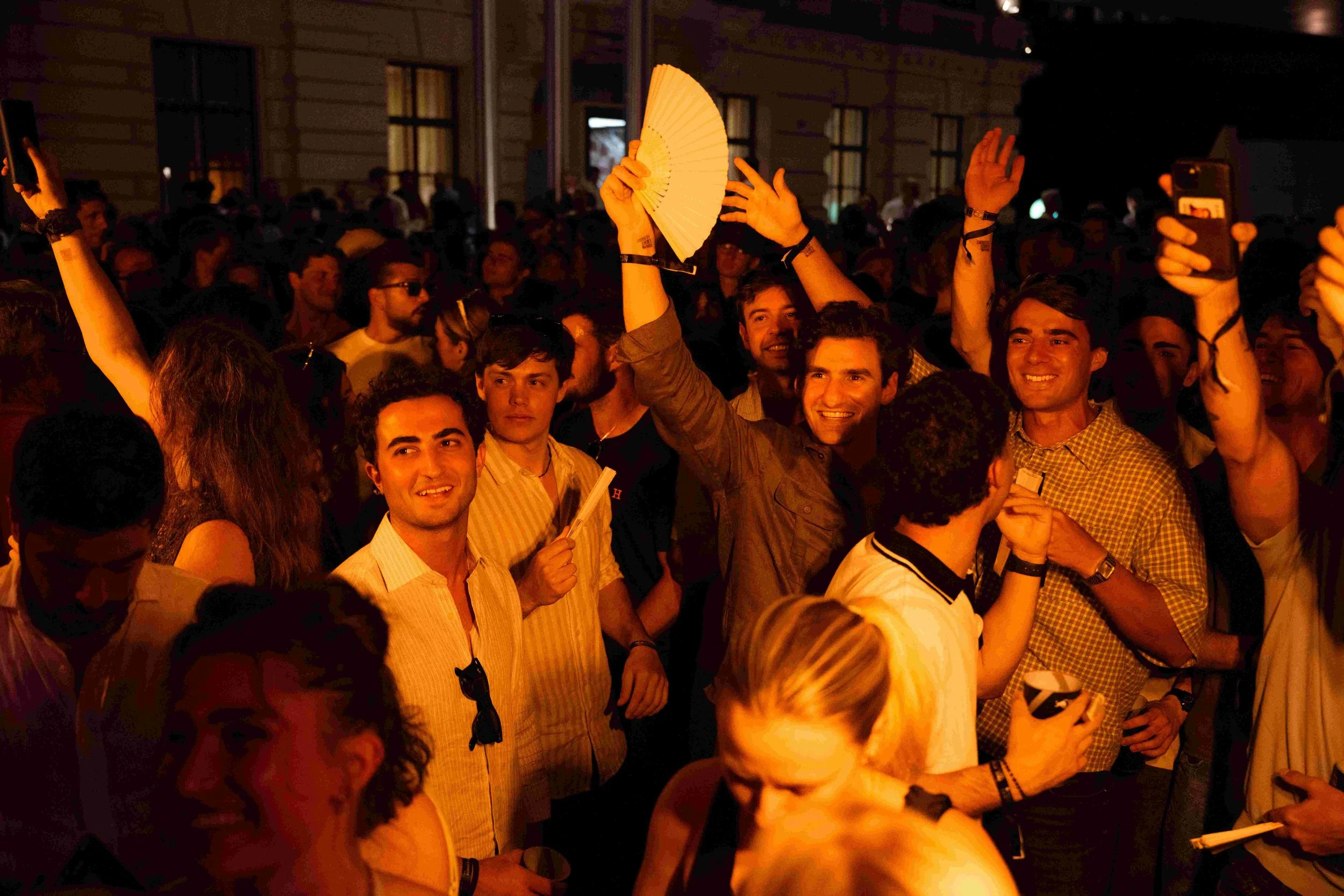 A large crowd of people attending a nighttime event, with many smiling and holding up phones. One person in the center is holding a white fan above their head. The background shows a building with lit windows.