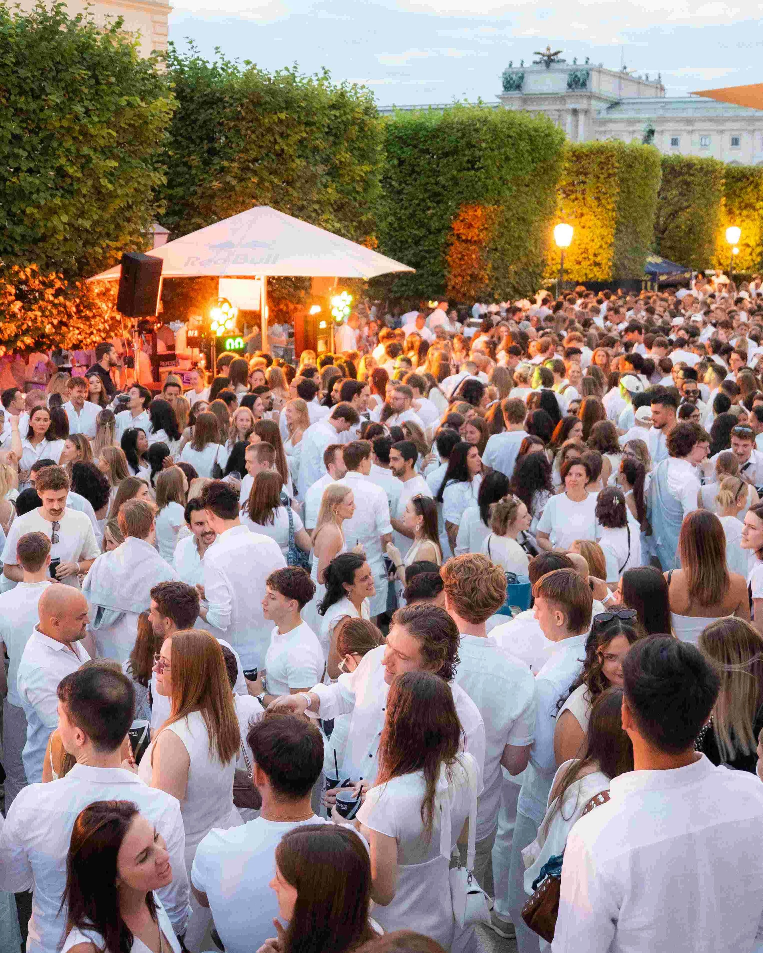 Crowd of people dressed in white in an outdoor setting during the evening, with trees, street lights, and historic building in the background.