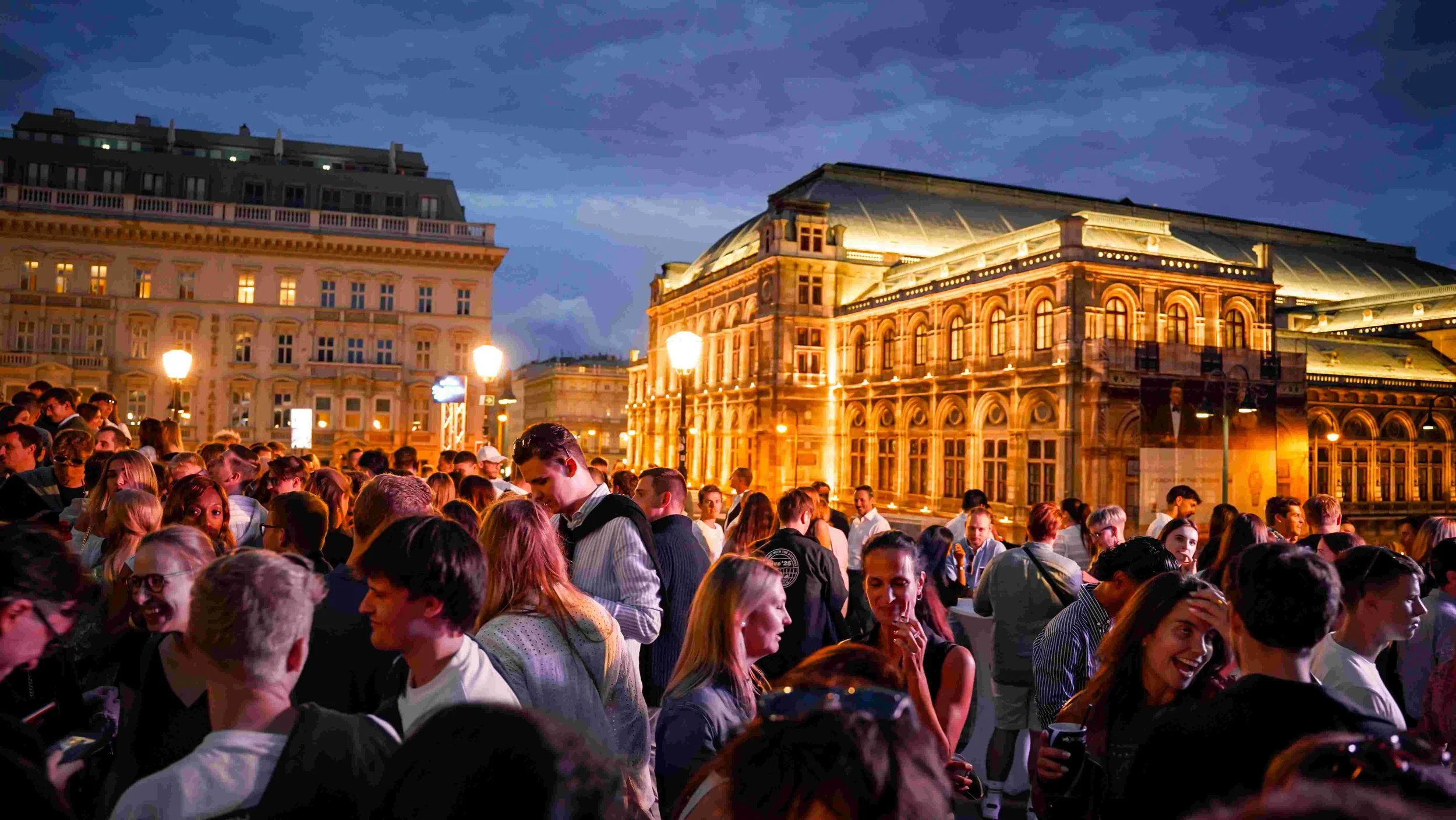 Crowd of people gathered outdoors in front of historic buildings illuminated at night.