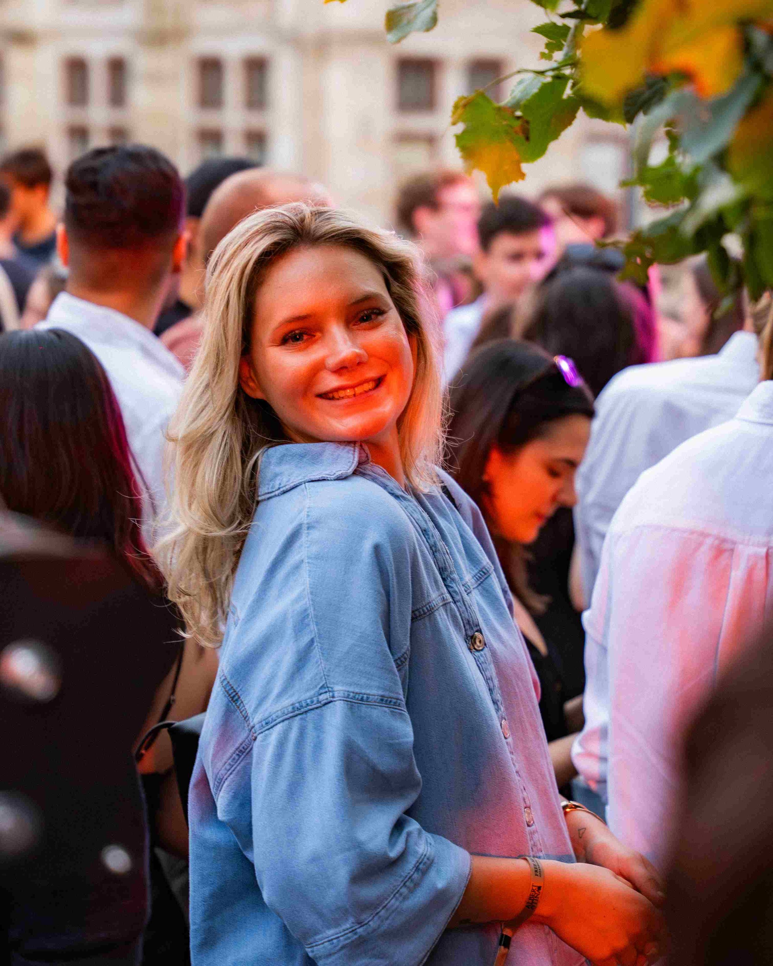 A group of people gathered outdoors during dusk, with a woman in a denim jacket smiling at the camera in the foreground. Others around her are engaged in conversation or looking elsewhere, with buildings visible in the background.