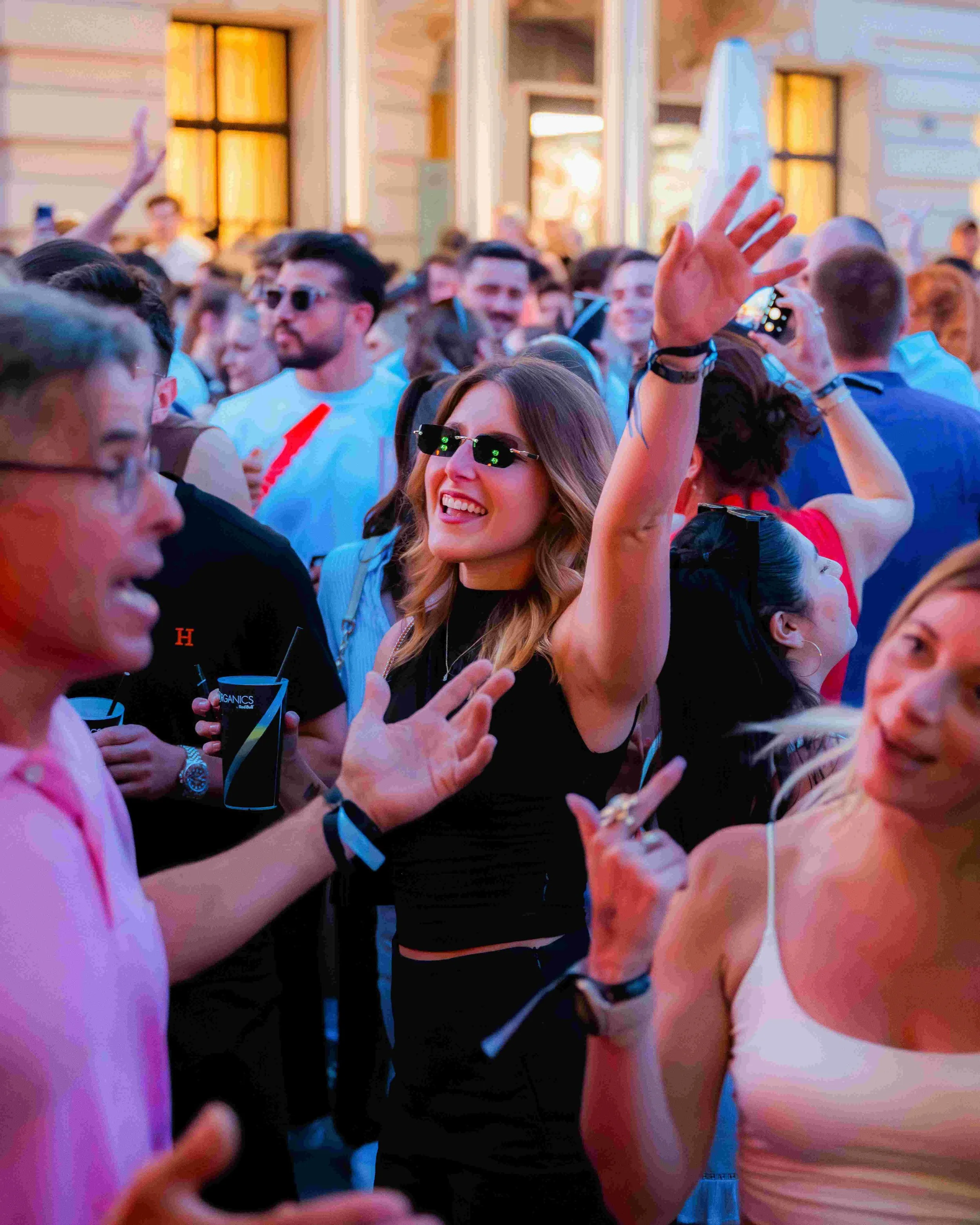 A group of people at a crowded indoor event, some wearing sunglasses and wristbands, with a woman smiling and raising her hand, and others engaging in conversation.