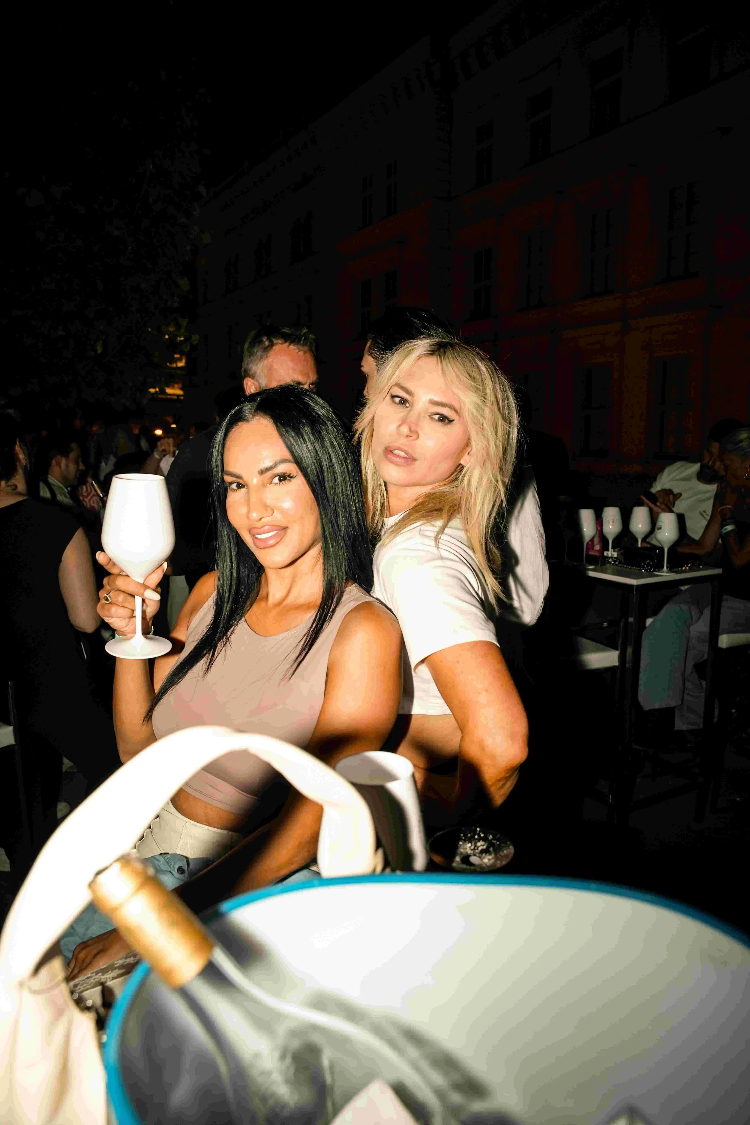 Two women at an outdoor event during the evening, one with black hair holding a wine glass, and the other with blonde hair, posing for the camera.
