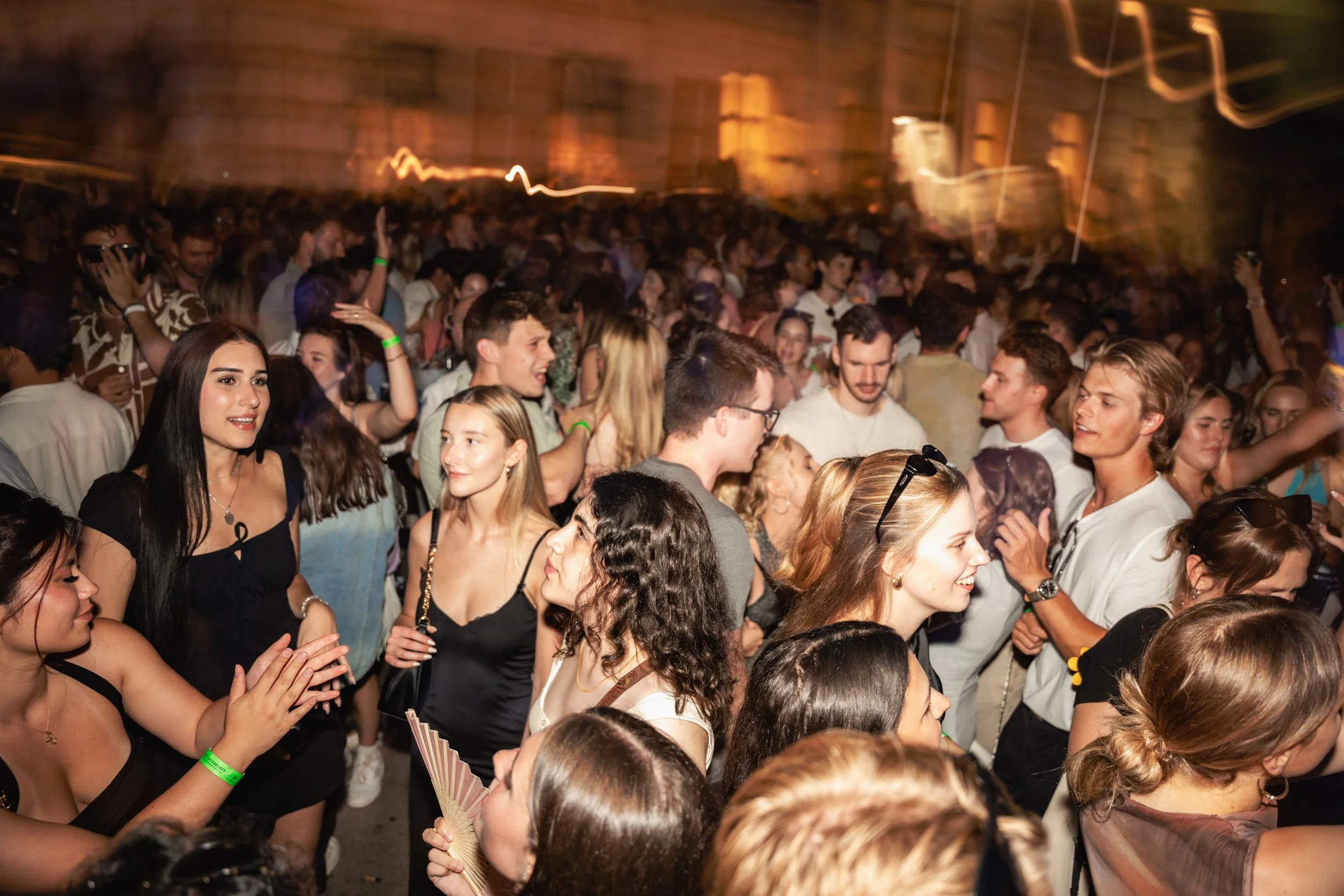 Crowd of young people gathered at a lively event or party, many smiling and talking with each other, some wearing sunglasses and wristbands, with warm lighting in the background.