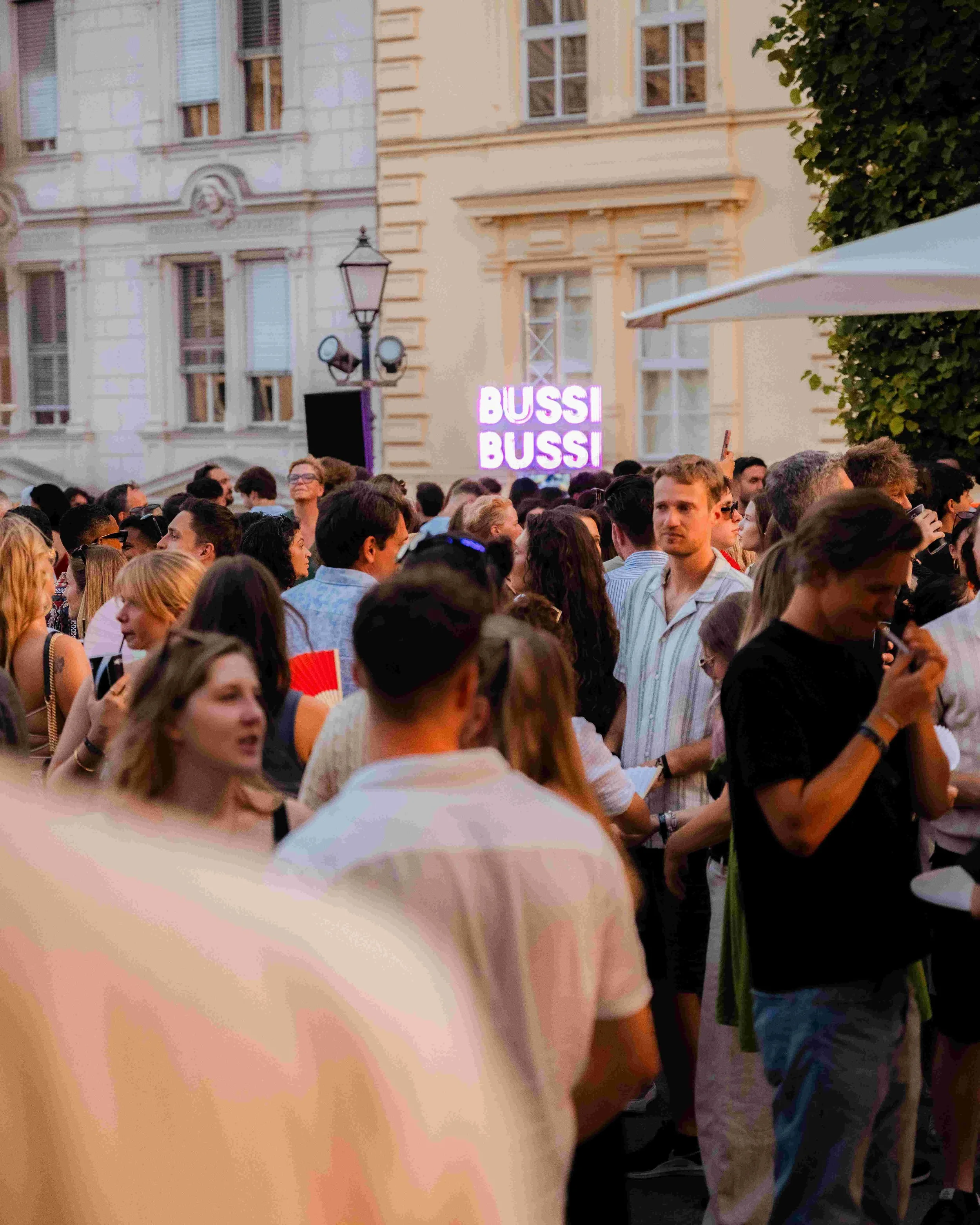 A large crowd of people gathered outdoors in front of buildings with European architectural style, including large windows and ornate details. There is a sign that reads 'BUSSI BUSSI' in the background, and some people are looking at their phones whi