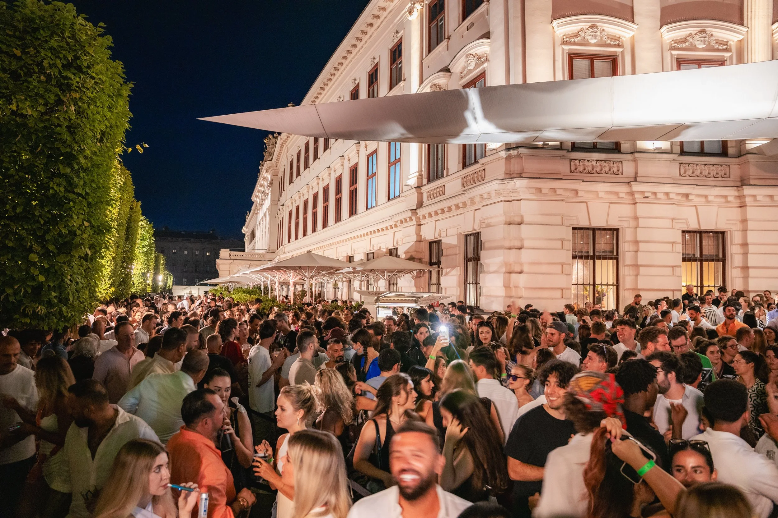 Nighttime outdoor gathering next to a historic building with large windows, lit up, with a crowd of people socializing, some taking photos, and a large modern art sculpture extending from the building.