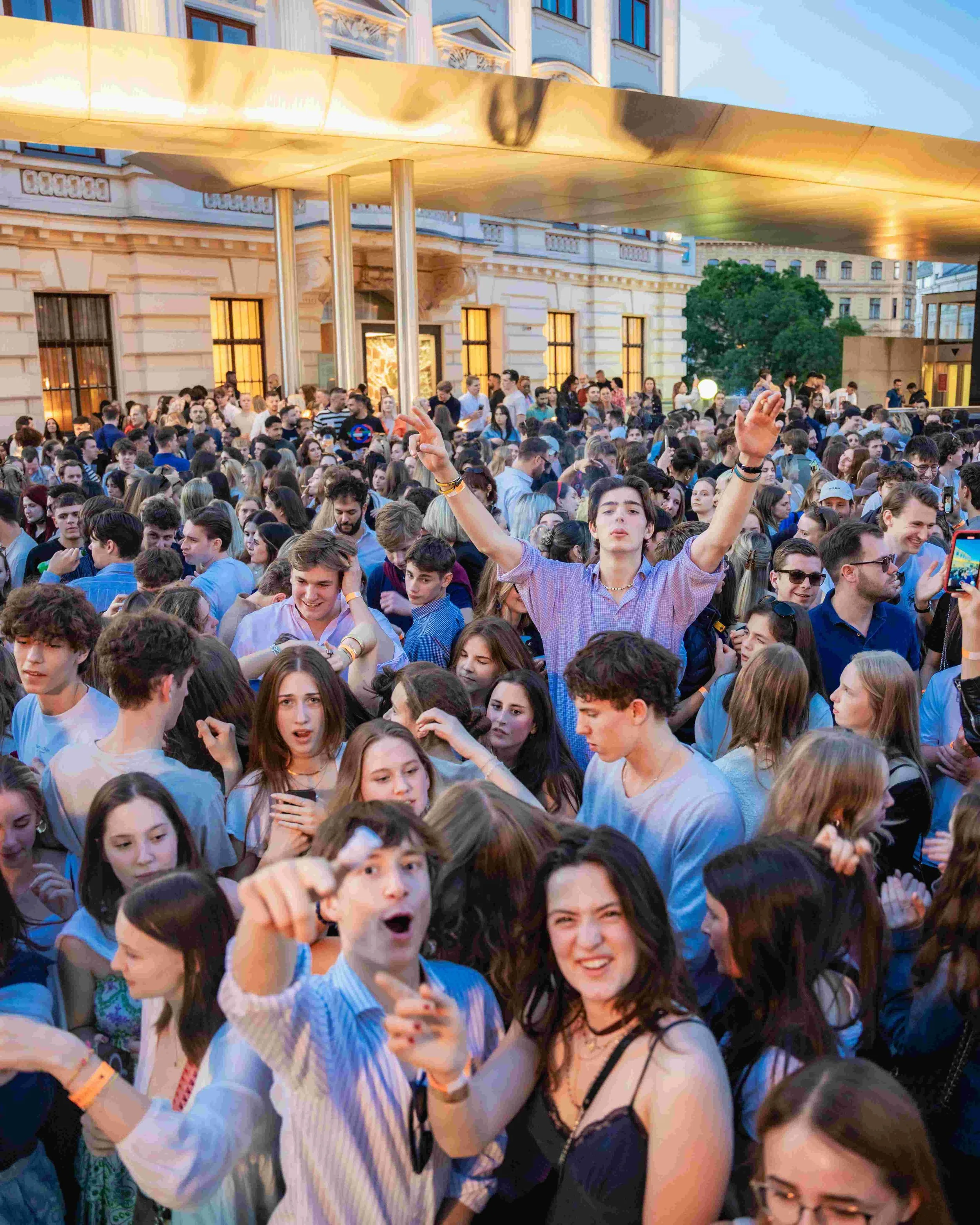 Crowd of young people at an outdoor event under a gold-colored canopy, with a historic building in the background.