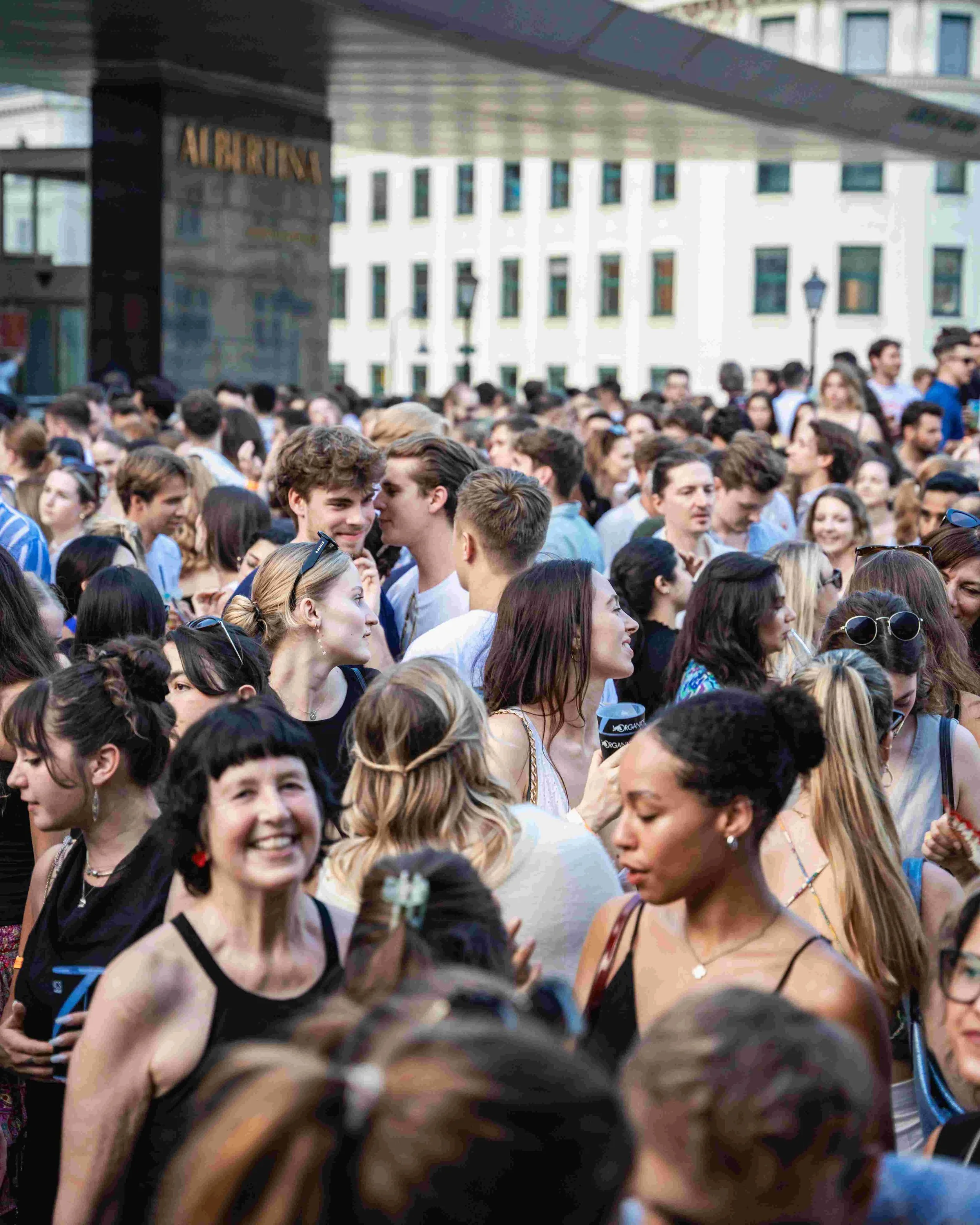 Crowd of young people gathered outside a modern building with a sign that reads 'ALBERTINA,' some smiling and chatting in an urban setting during daytime.