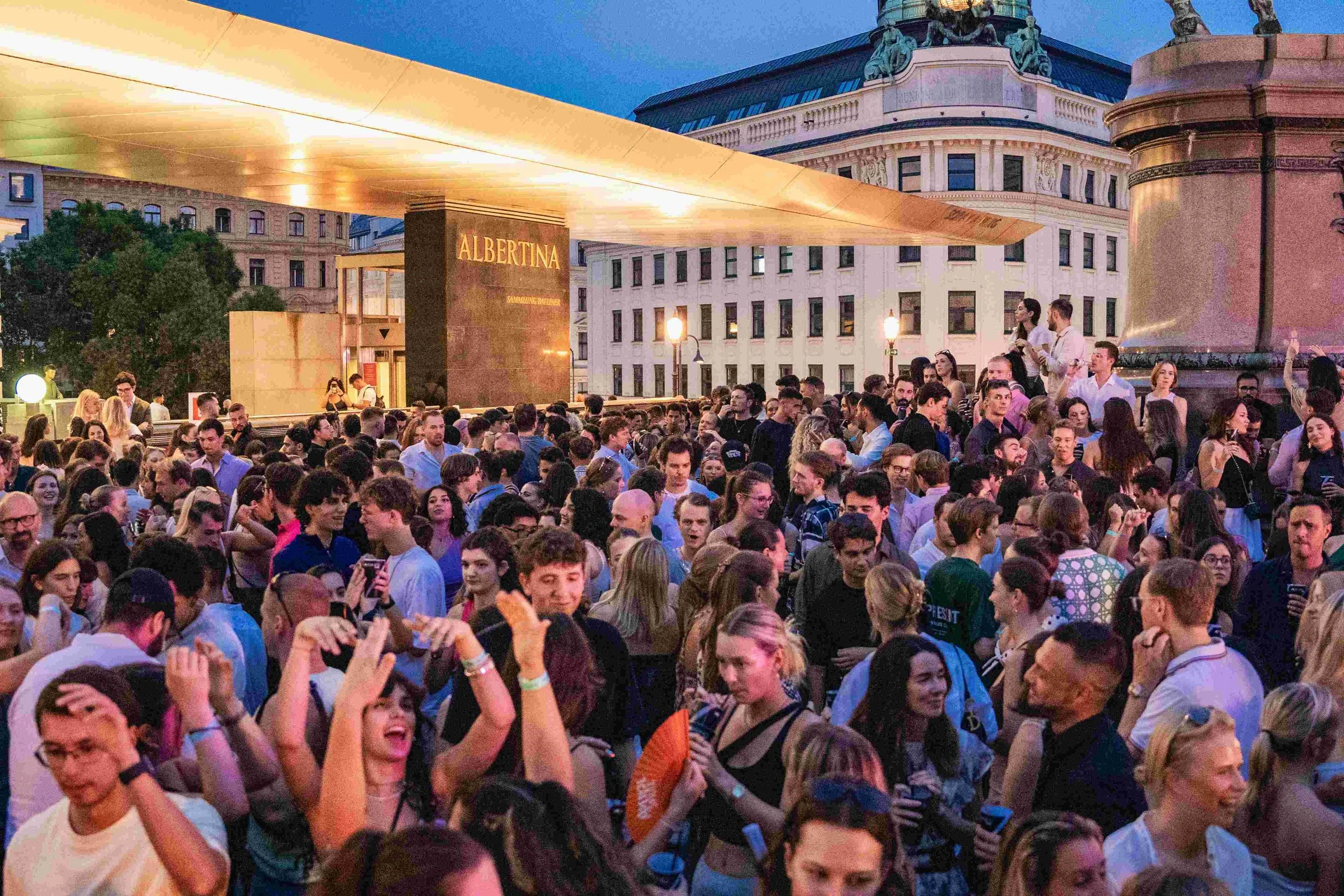 A large crowd of people gathered outdoors in front of a building with a sign that reads "ALBERTINA." The scene is during dusk or early evening with a clear blue sky. The people are engaging in conversation, taking photos, and enjoying the event.