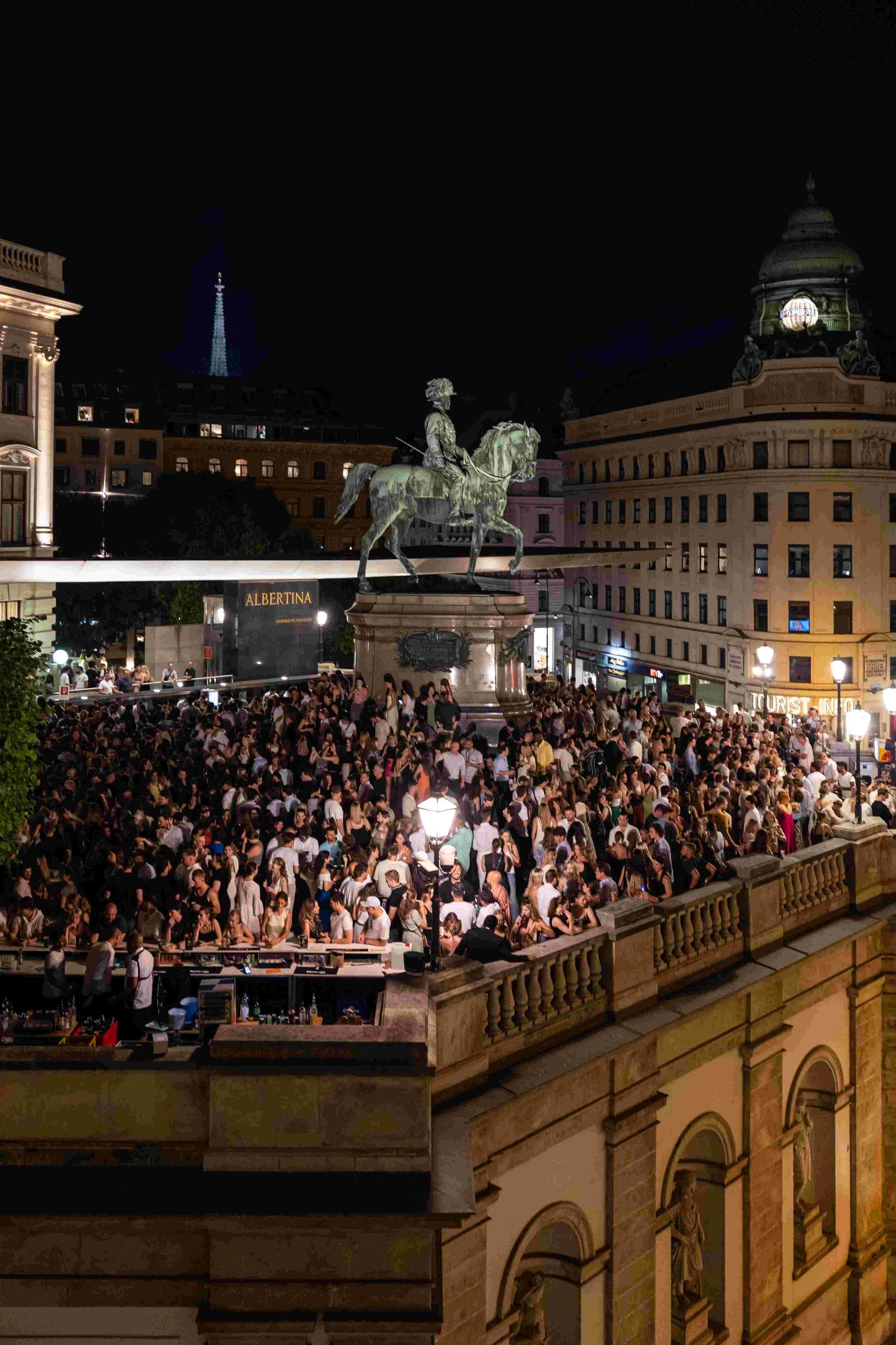 Nighttime scene of a busy city square with a large crowd and a statue of a man on horseback in the center. The statue is elevated on a platform. Surrounding buildings have decorative architecture, and there is a historic building with statues on the 