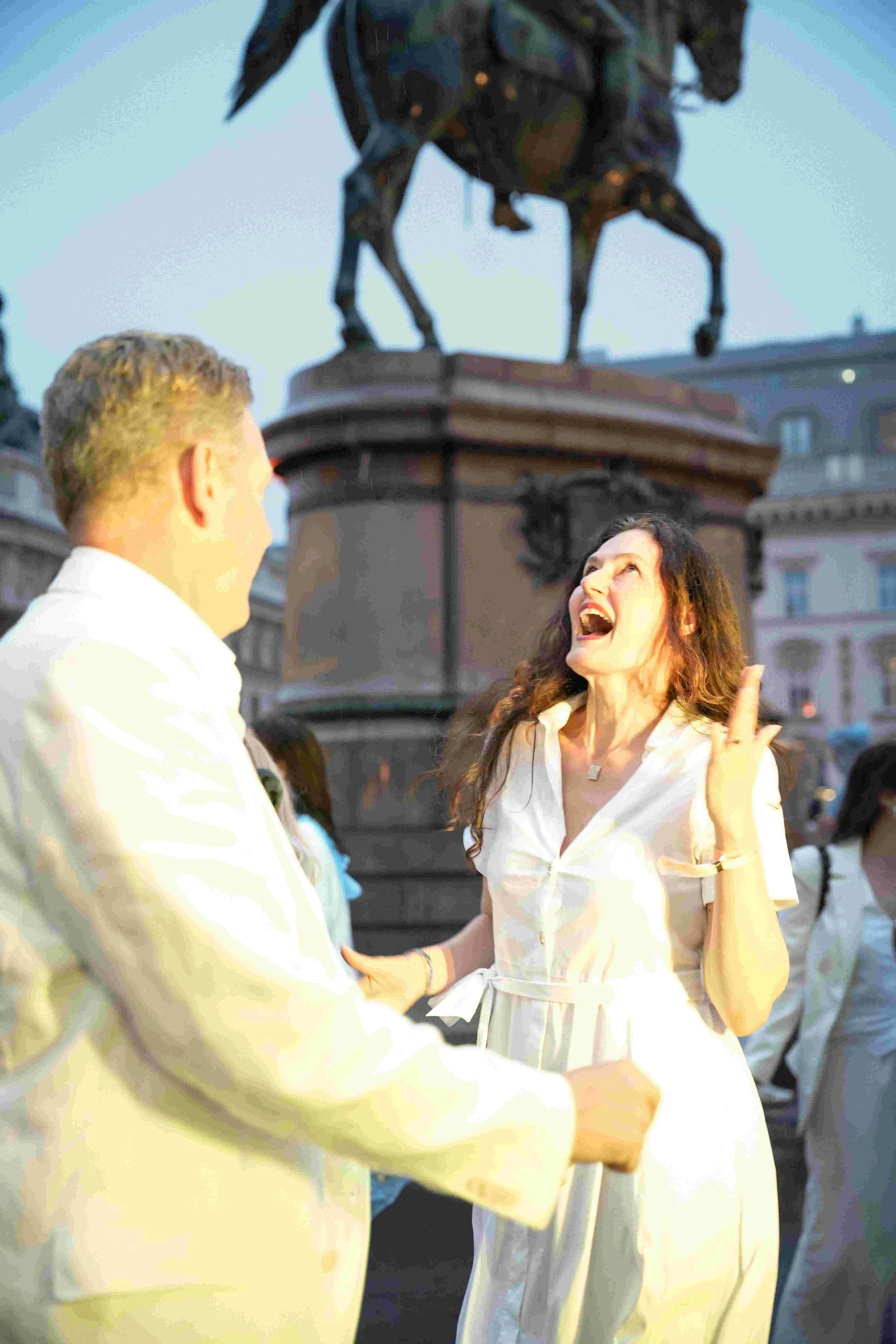Woman with long brown hair, wearing a white dress, talking and gesturing with her arm raised, standing in front of a man with short blonde hair, in an outdoor setting with a large statue of a horse and rider in the background, and other people around